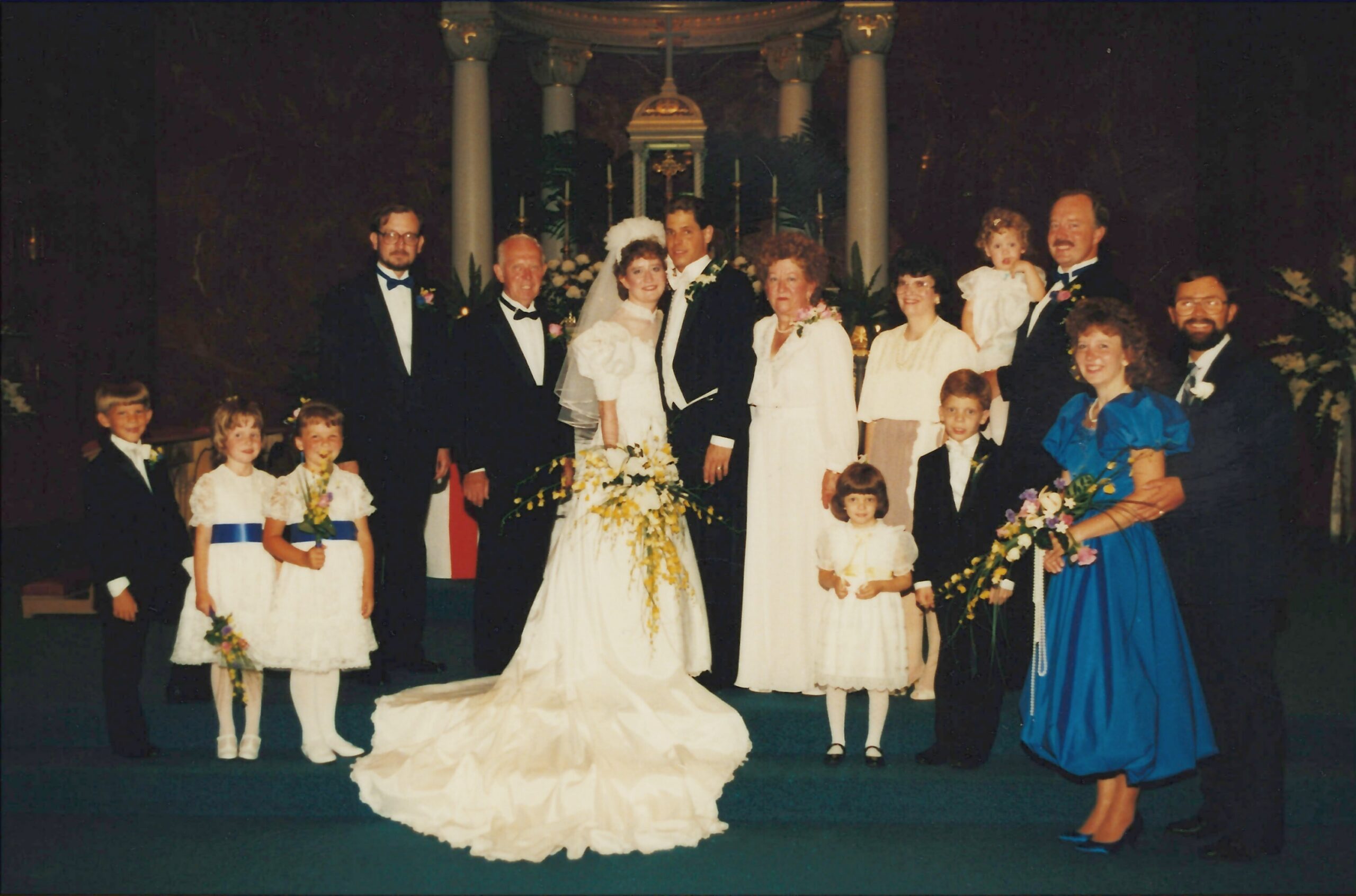 Bride and groom at center posing with family and wedding party inside a church altar area, wearing formal attire.