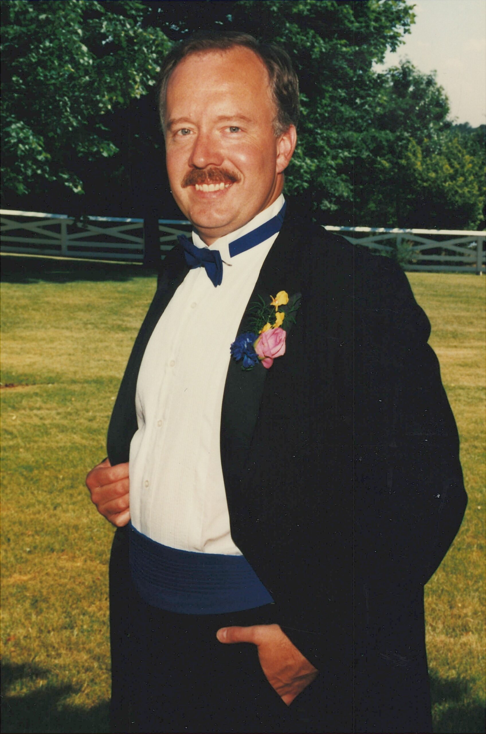 Man in a black tuxedo and blue bow tie posing outdoors with a pink rose boutonniere, smiling at the camera.