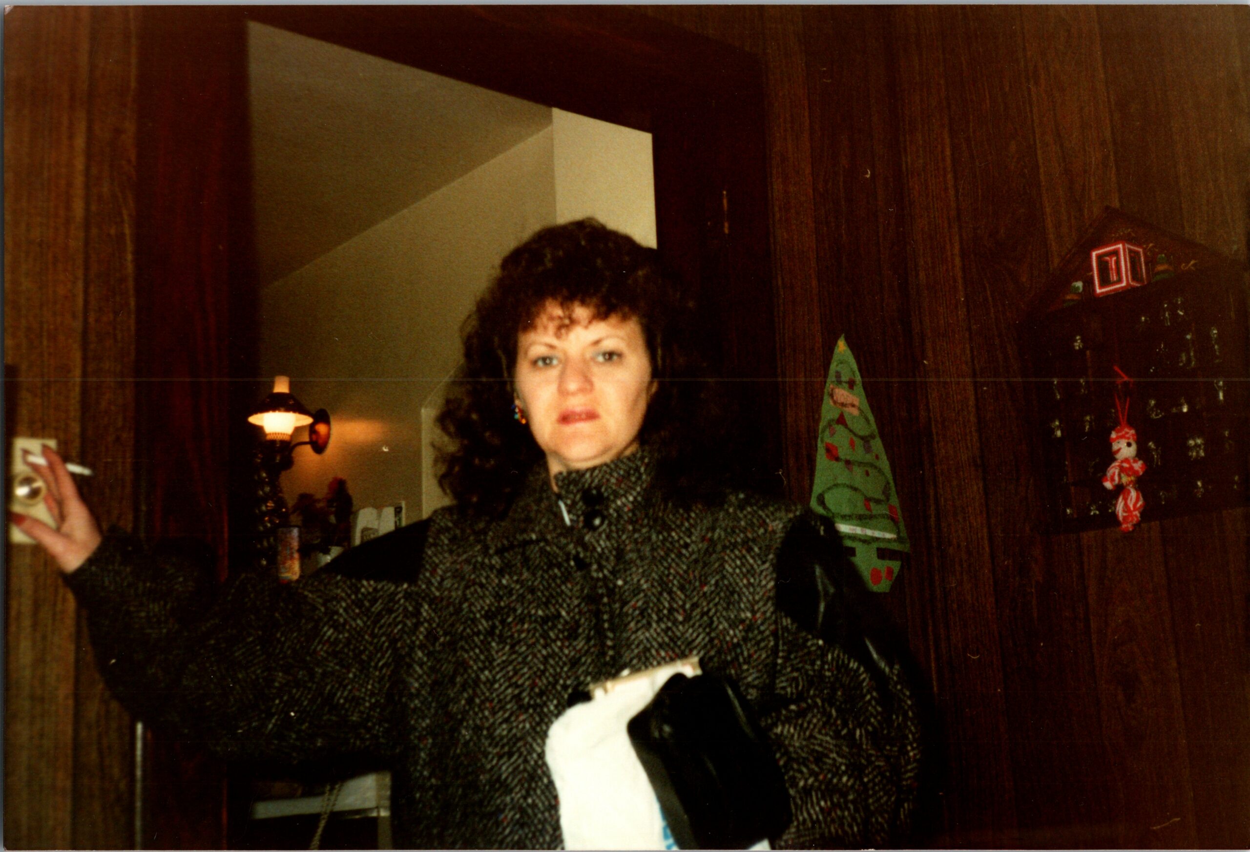 Woman with dark curly hair wearing a textured coat, standing indoors among Christmas decorations on wood-paneled walls.