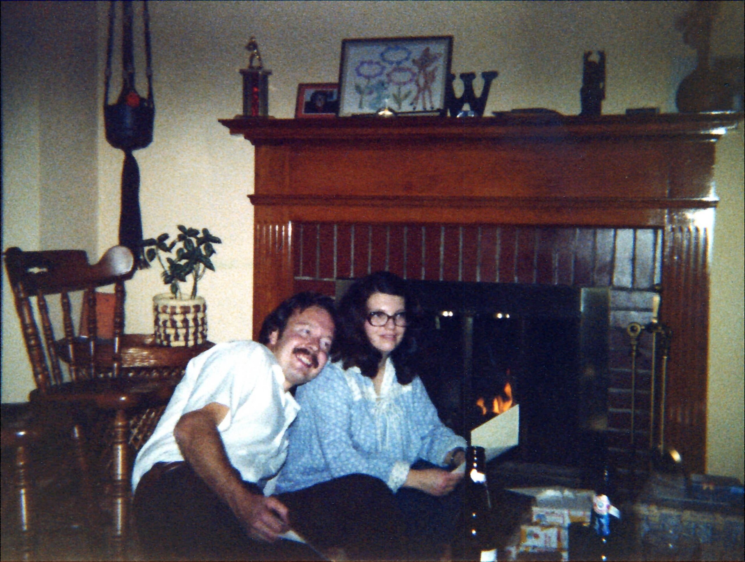 Three people sit and smile in a living room in front of a wooden fireplace mantle, with a lit fire to the right and decorative items (framed photo, blue 'W' letters) atop the mantel.