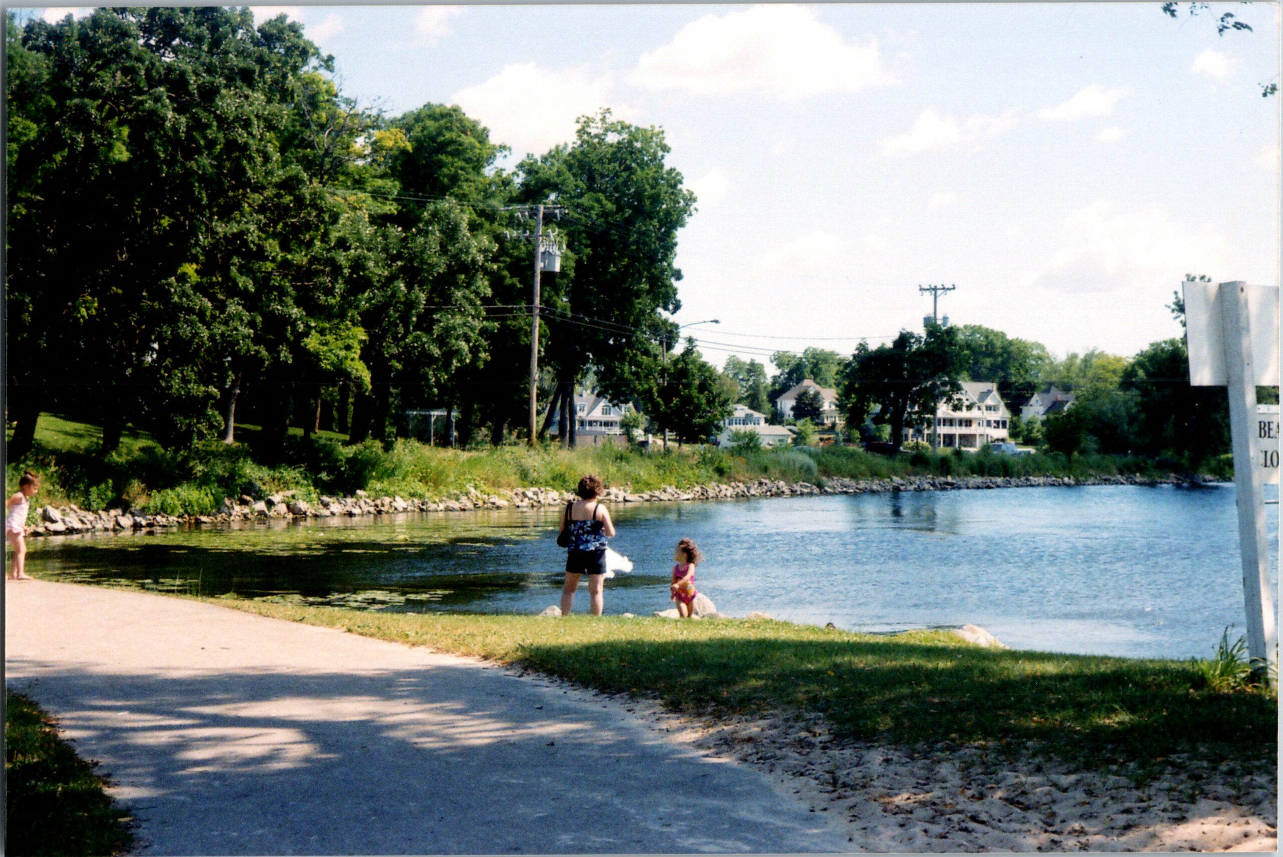 Families enjoy a riverside park: two children near the water with adults, trees lining the shore and houses in the background.