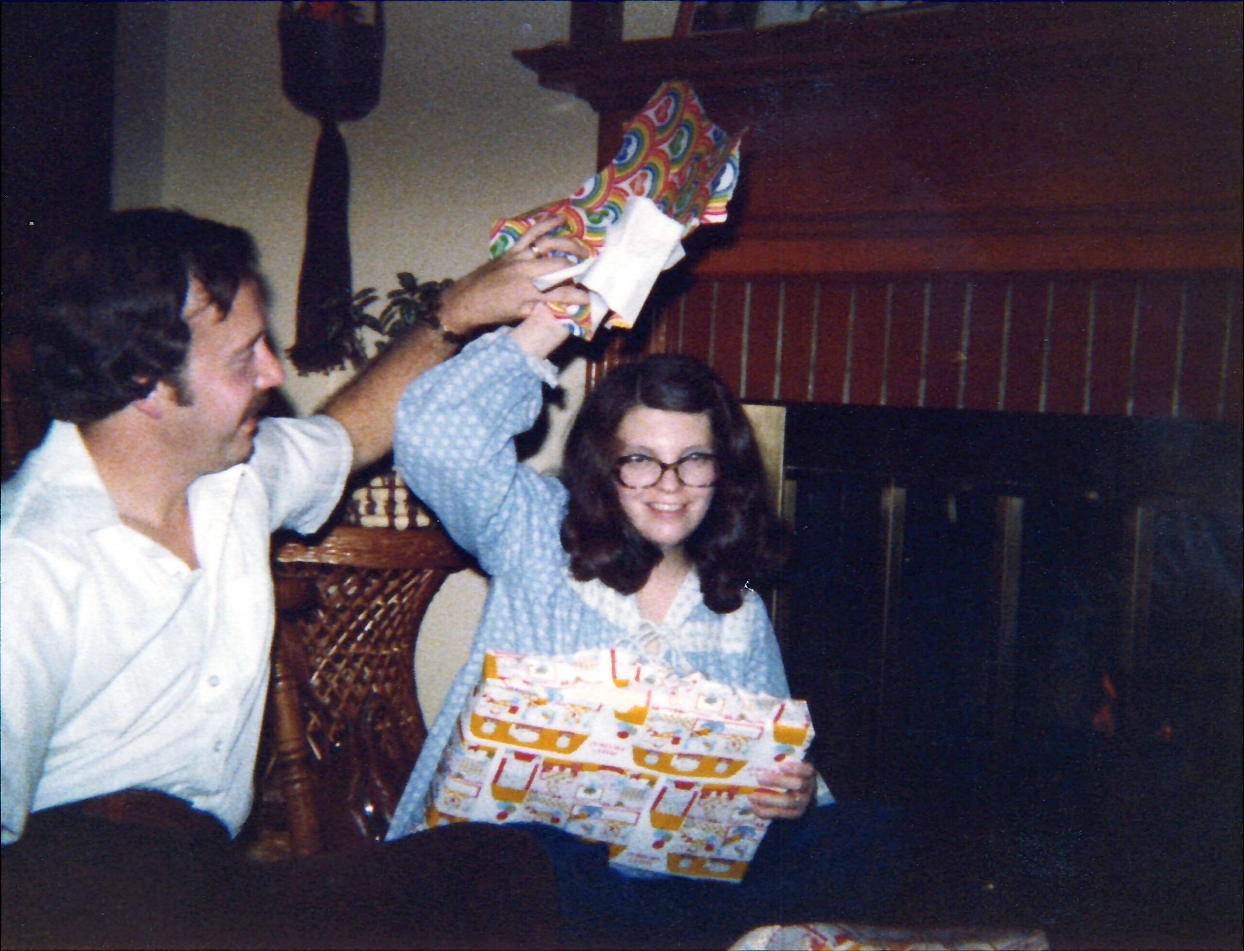 People at a small party celebrate as a woman raises a large colorful wrapped gift above her head.