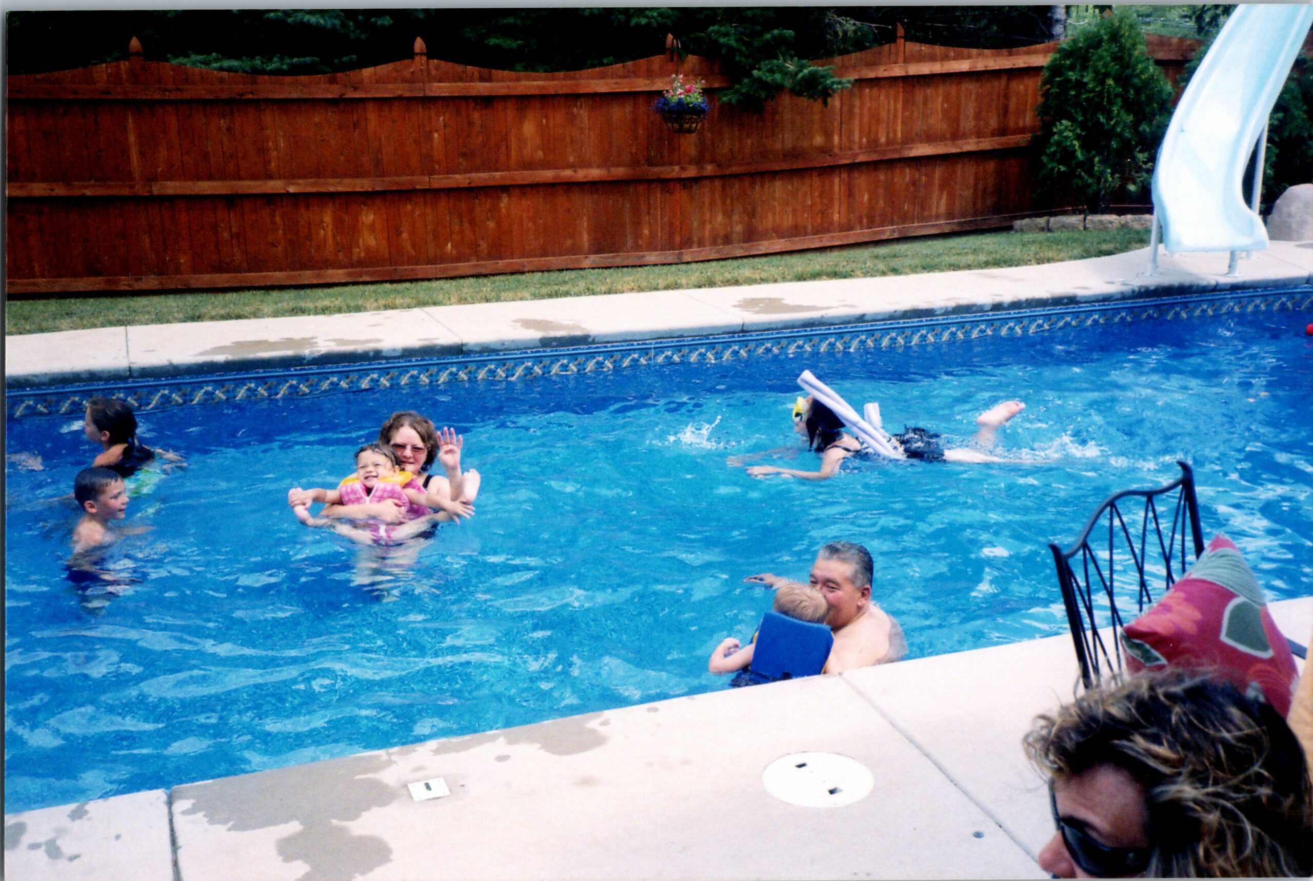 Family splashes in a backyard pool with a wooden fence and a blue slide in the background, kids wearing life jackets.