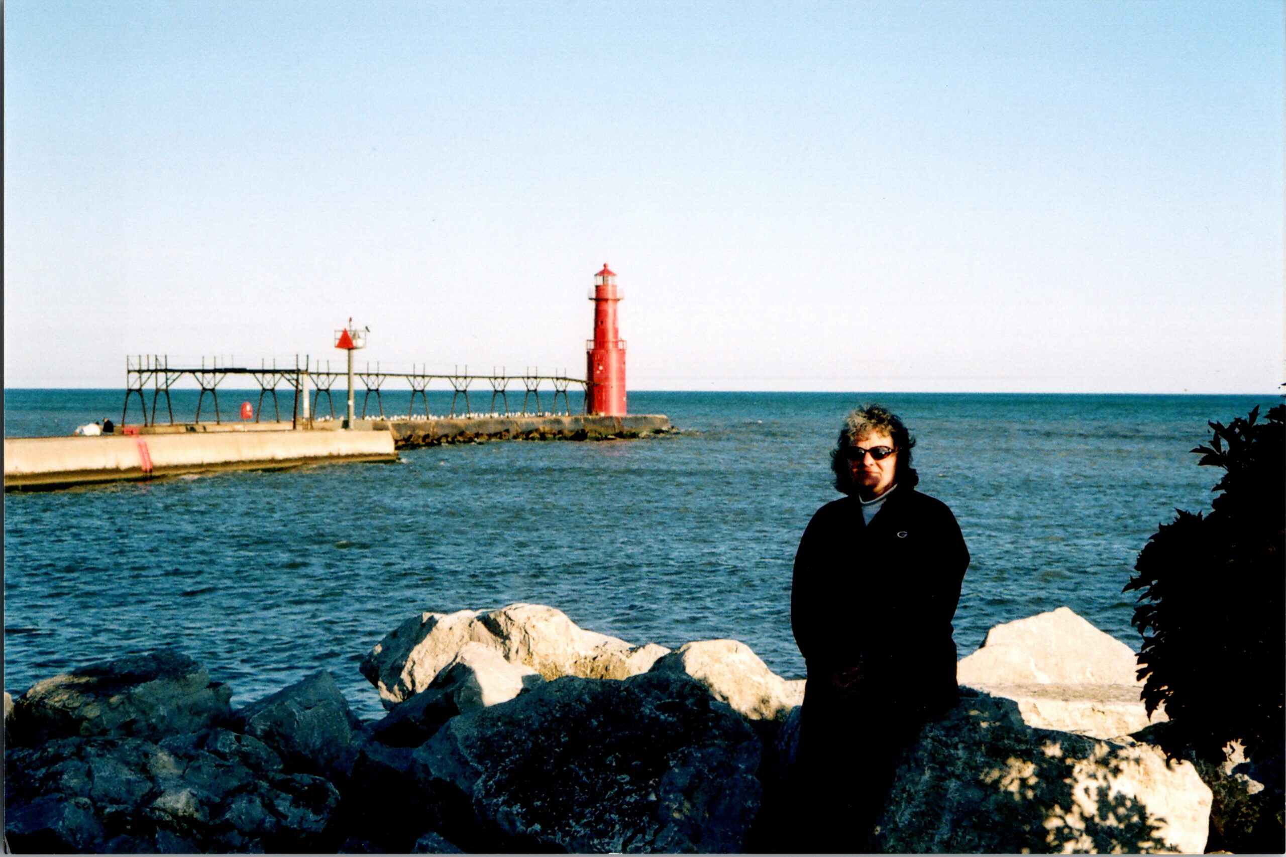 Red lighthouse on a jetty extending into blue water with a person in dark clothing and sunglasses standing on rocks in the foreground.