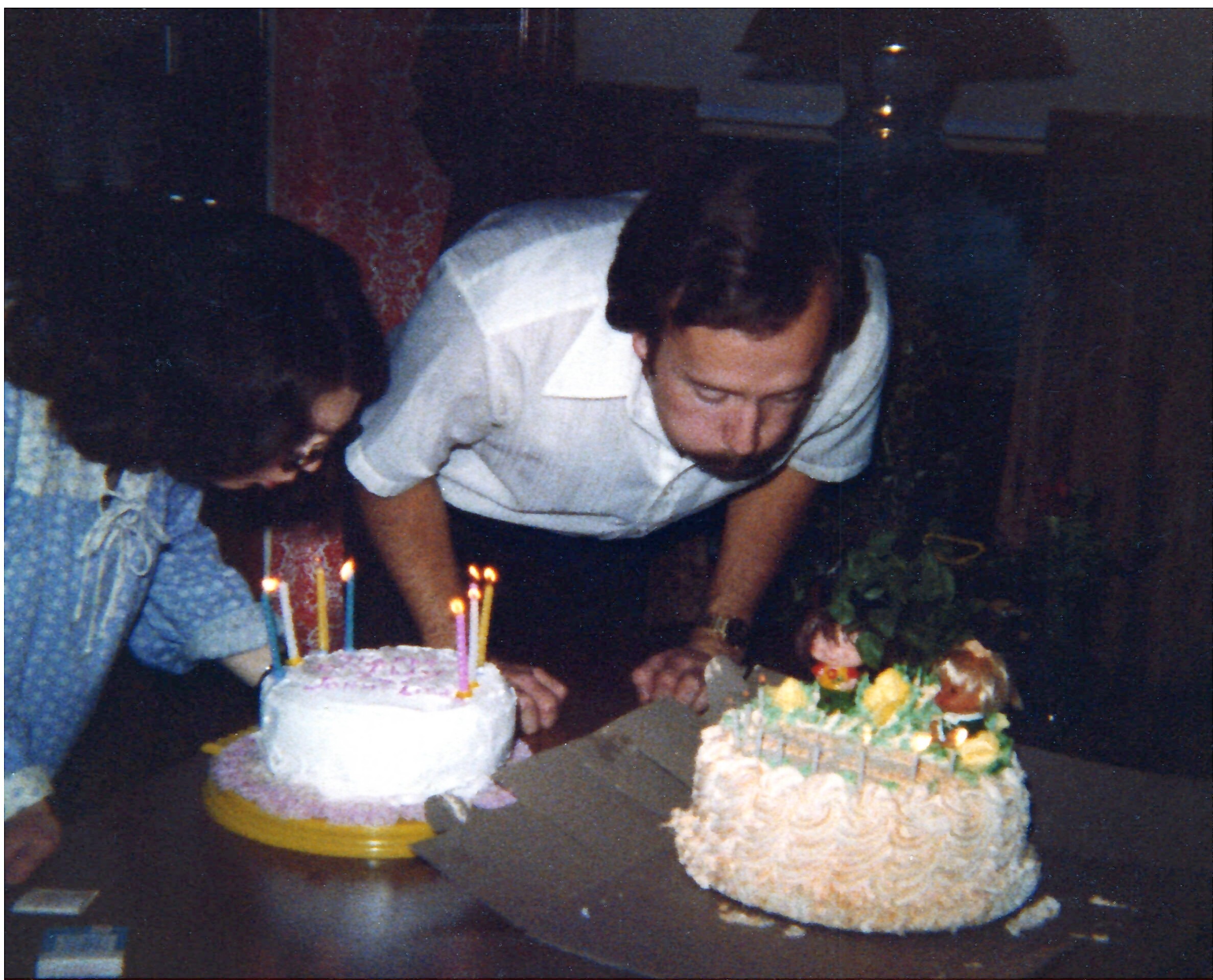 Two people lean over a table to cut a white frosted birthday cake with candles lit nearby.