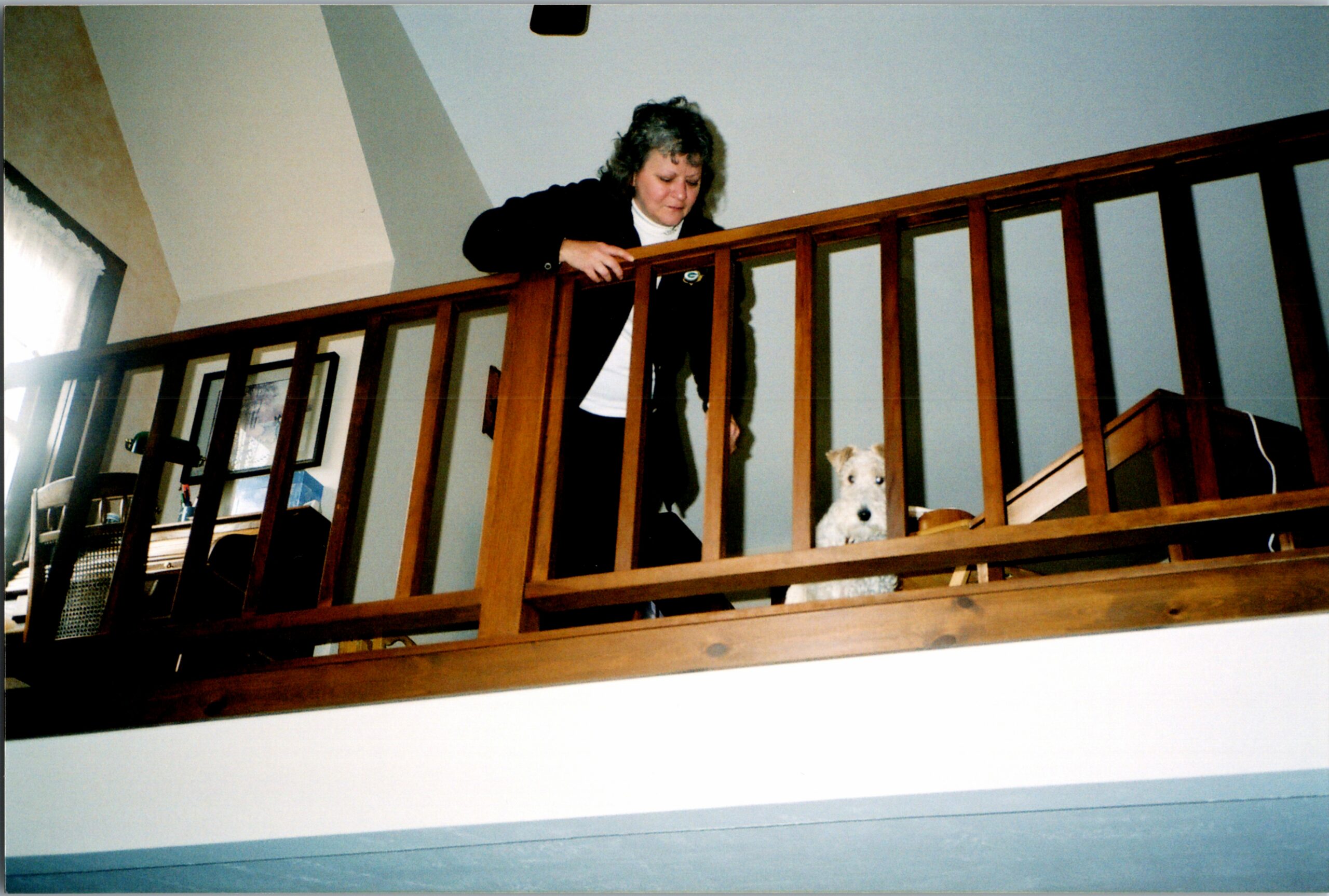 Woman leaning over a wooden railing on a loft, looking down at a white dog below the railing in a home interior