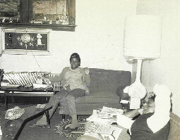 Black-and-white living room scene with two women seated: one on a sofa and another at a coffee table, surrounded by magazines, framed pictures on the wall, a tall floor lamp, and a low table.