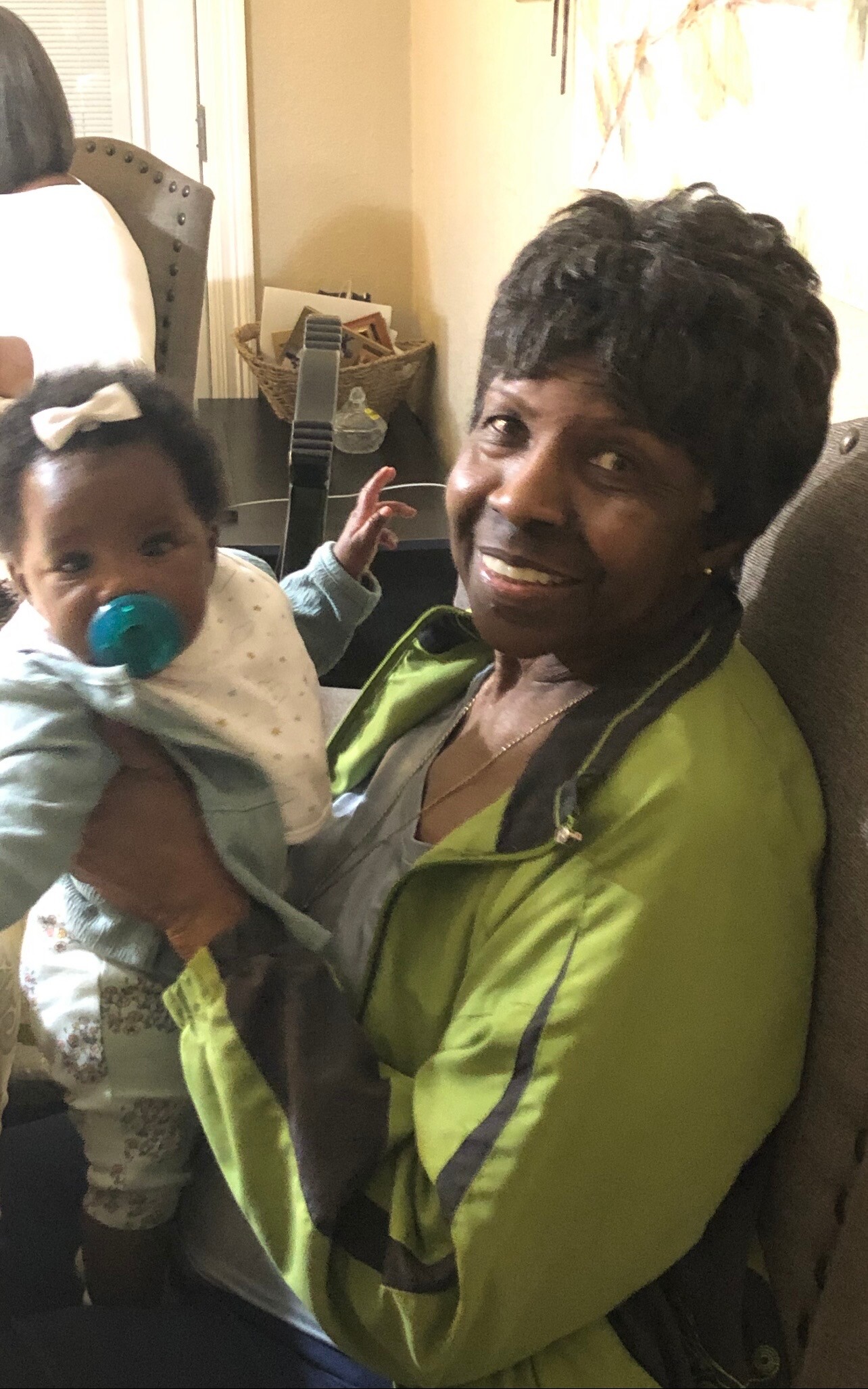Smiling woman in a green jacket holding a baby with a blue pacifier indoors.