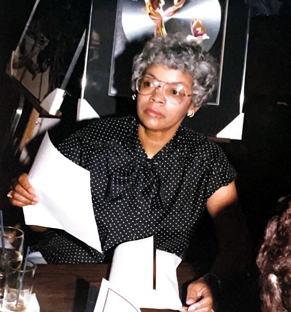 Older woman with gray curly hair and large glasses, wearing a black polka-dot blouse, holding papers at a table in a dimly lit event space.