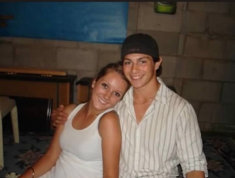 Young couple sitting closely, smiling at the camera; woman in a white tank top, man in a striped shirt and black cap.
