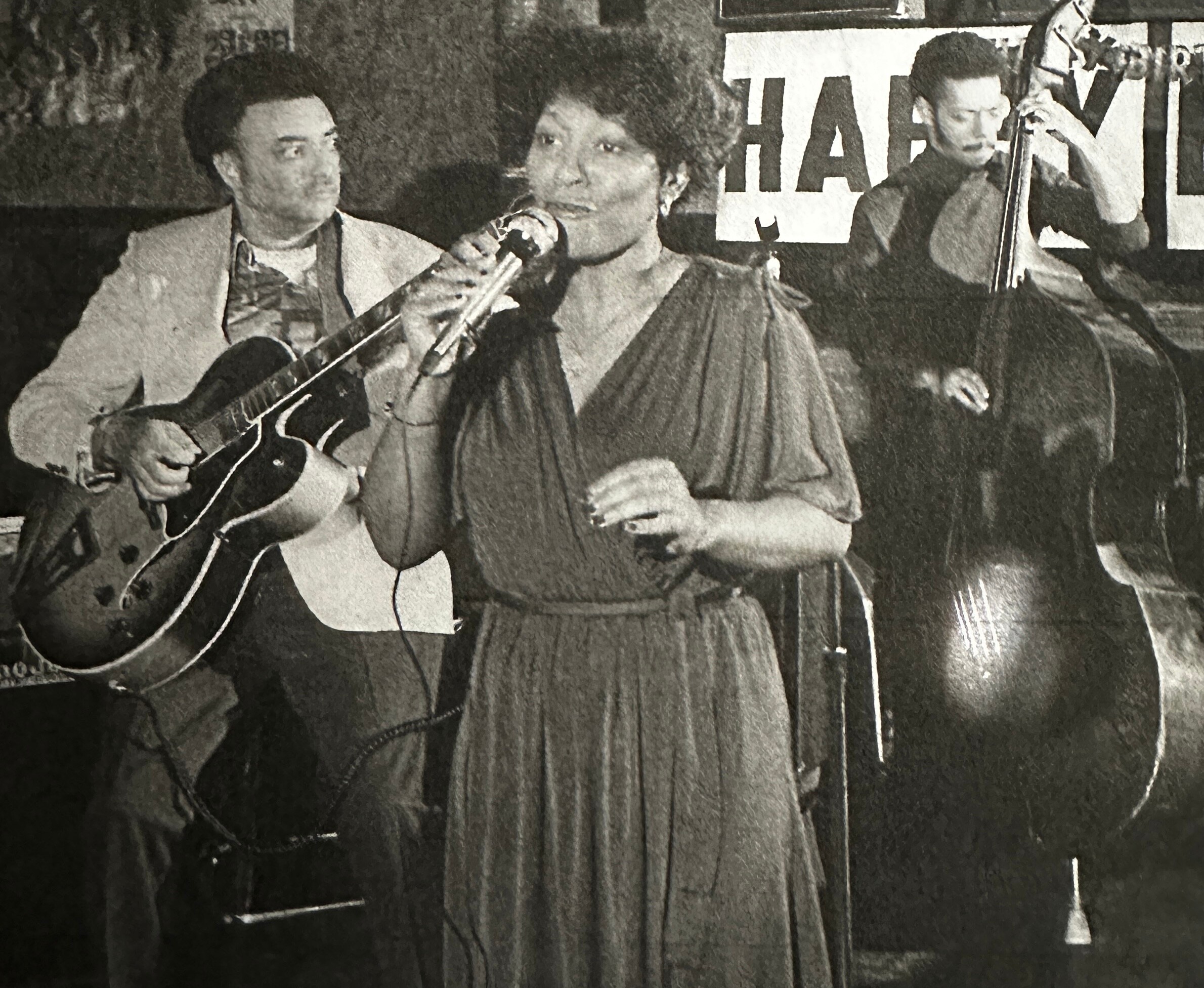 Black-and-white photo of a trio performing on stage: guitarist on the left, a female vocalist center, and a horn player on the right.