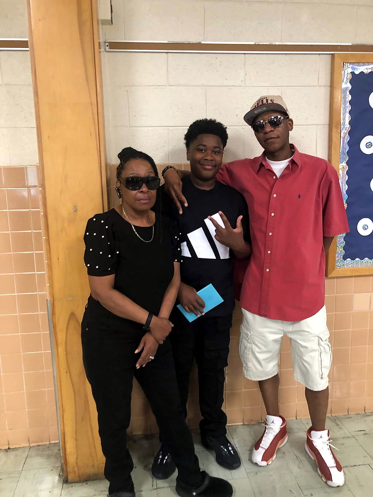 Three people pose in a school hallway: an older woman in black with sunglasses, a boy in a black graphic tee holding a blue card, and a tall teen in a red shirt and cap with sunglasses.