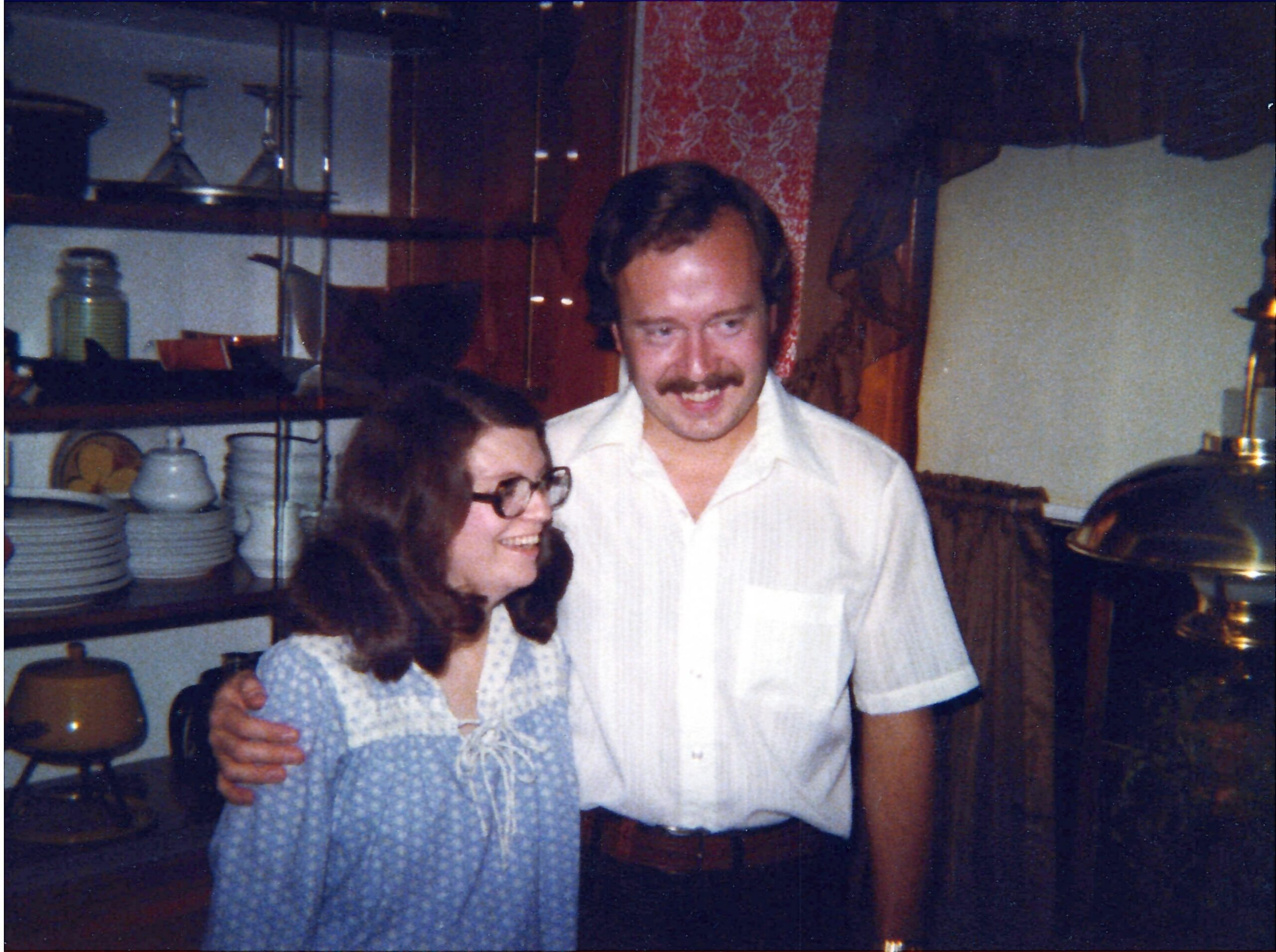 Smiling couple posing together in a home kitchen, with dishes and shelves in the background.