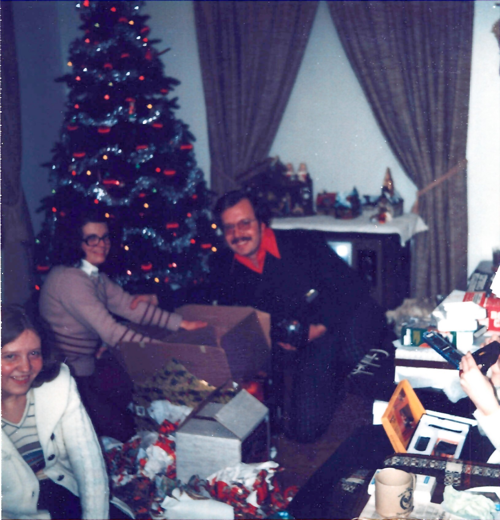 Family gathers around a decorated Christmas tree opening presents; colorful lights and gifts on the table and floor behind them.