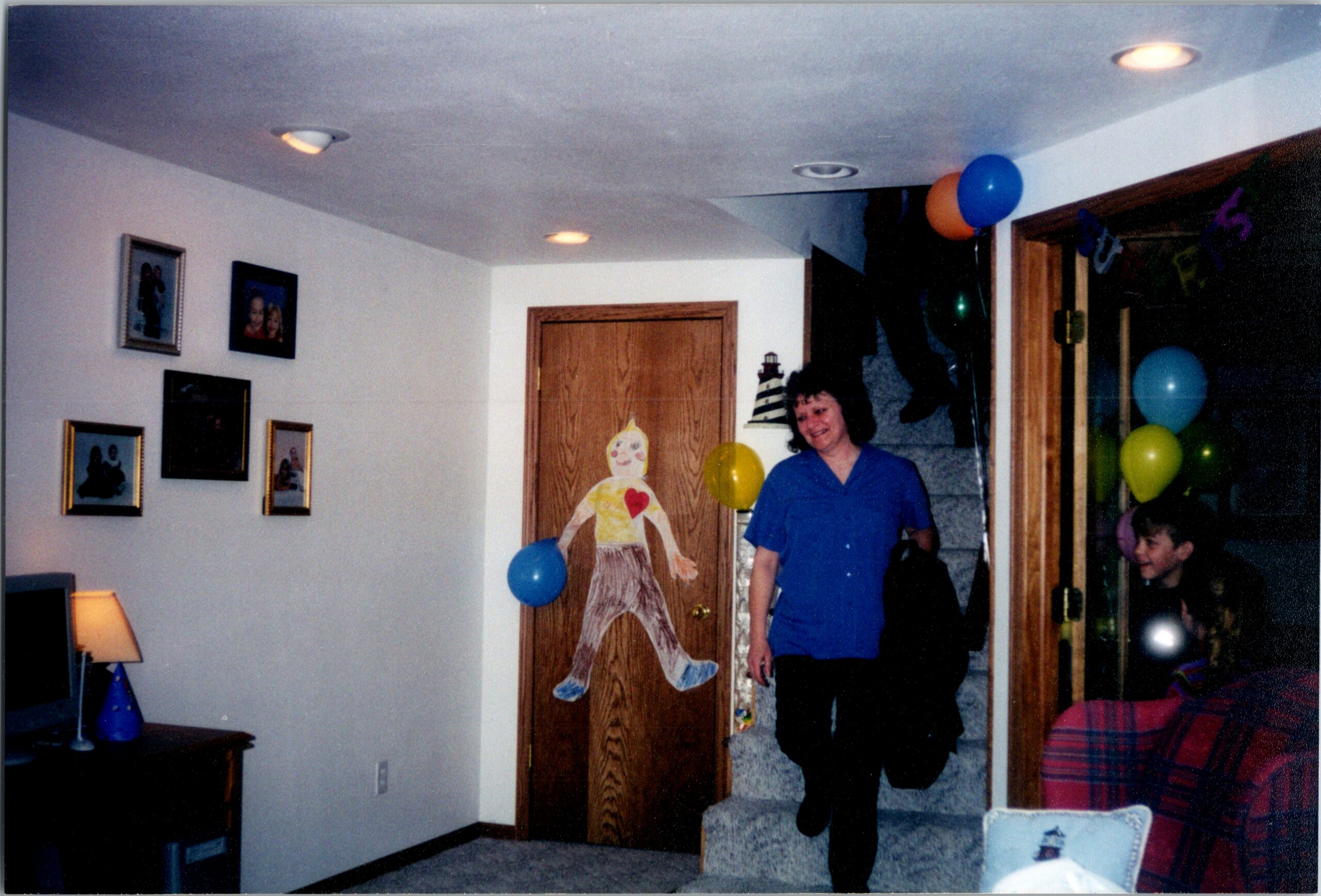 Woman in a blue shirt walks through a doorway in a home decorated with balloons for a party.parked by a paper cutout figure on the door and family photos on the wall.