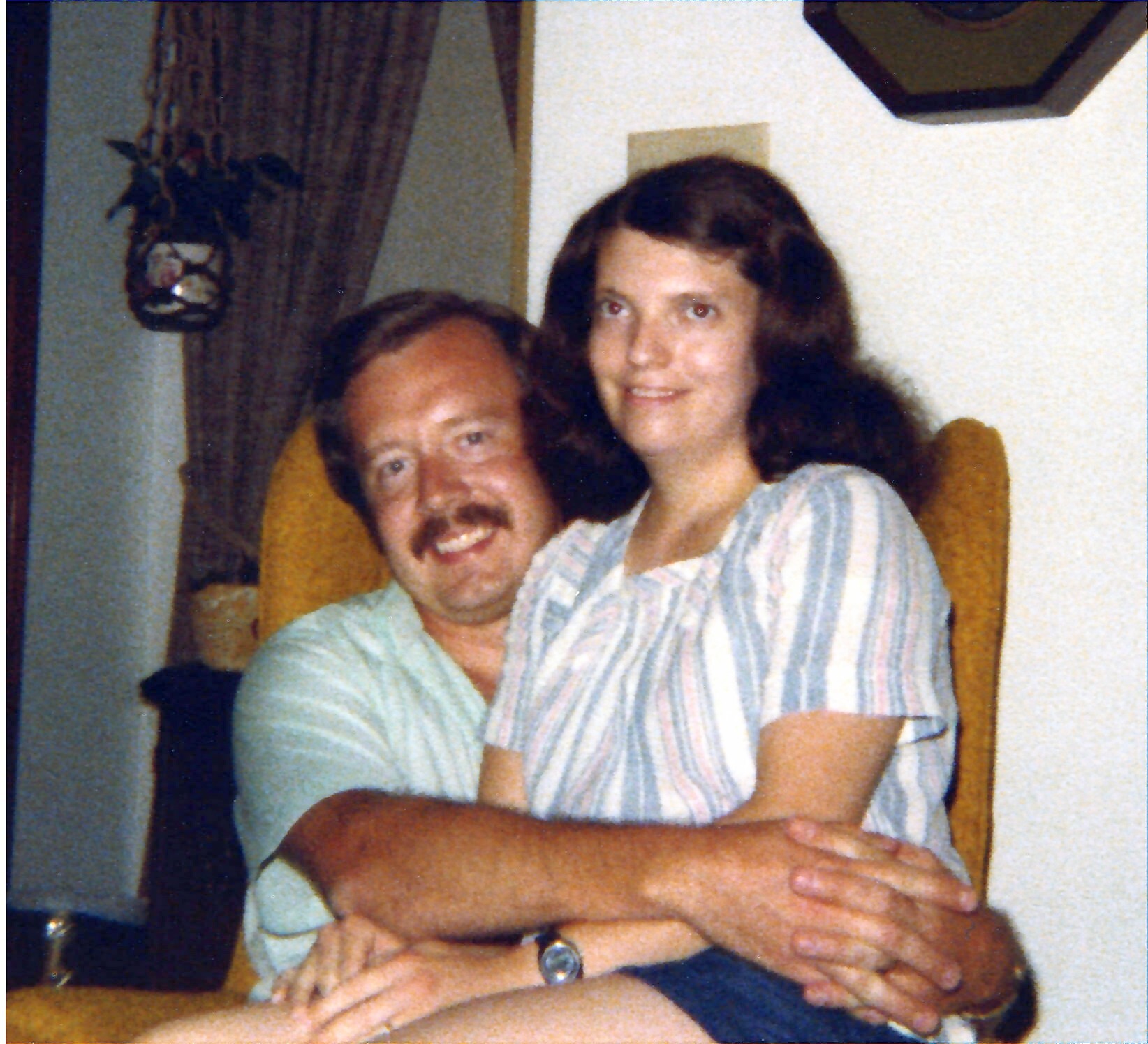 A man with a mustache hugs a smiling woman from behind as they sit close together indoors, arms resting on a table.