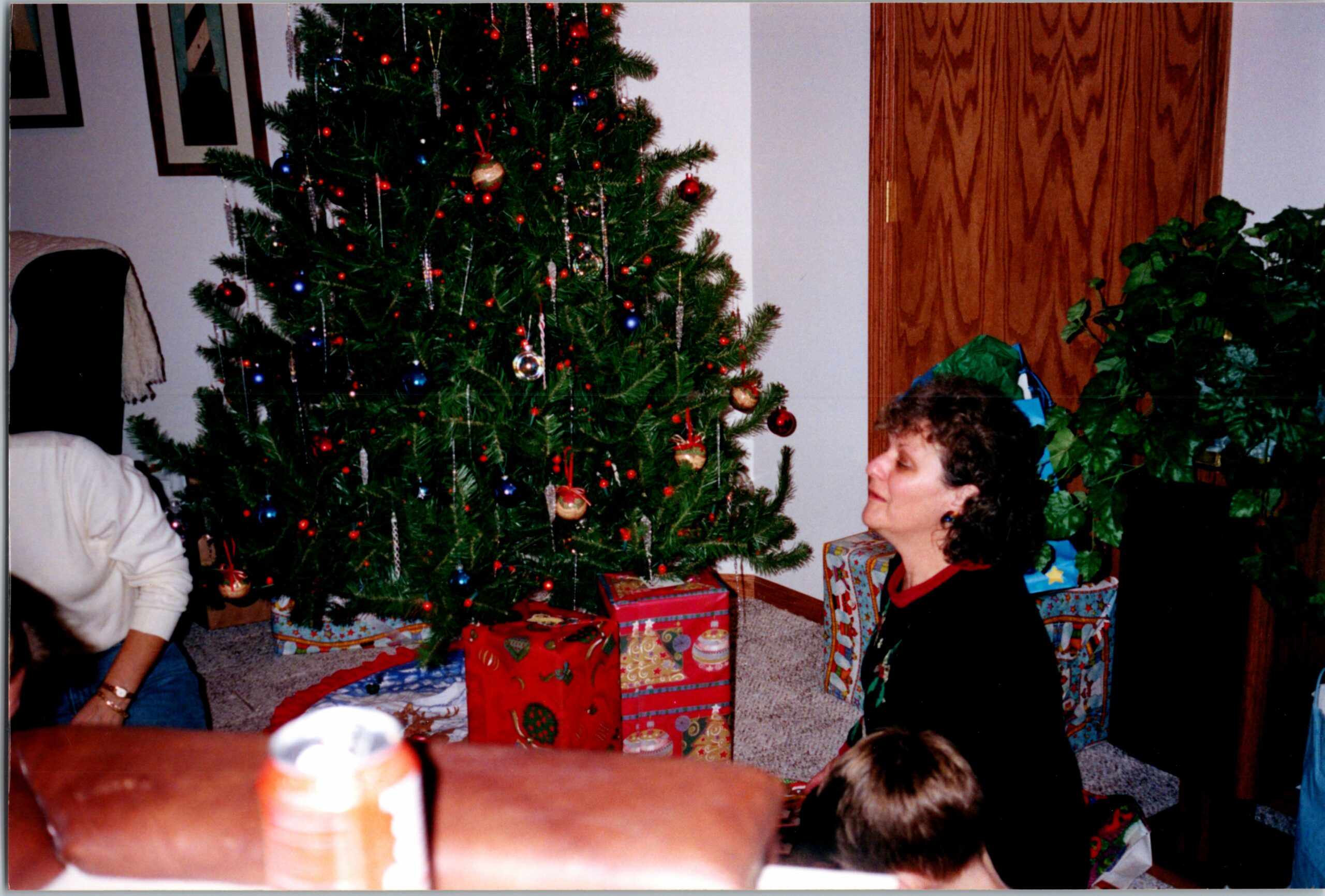 Christmas tree with red and blue ornaments and presents beneath; woman seated nearby in a domestic living room.