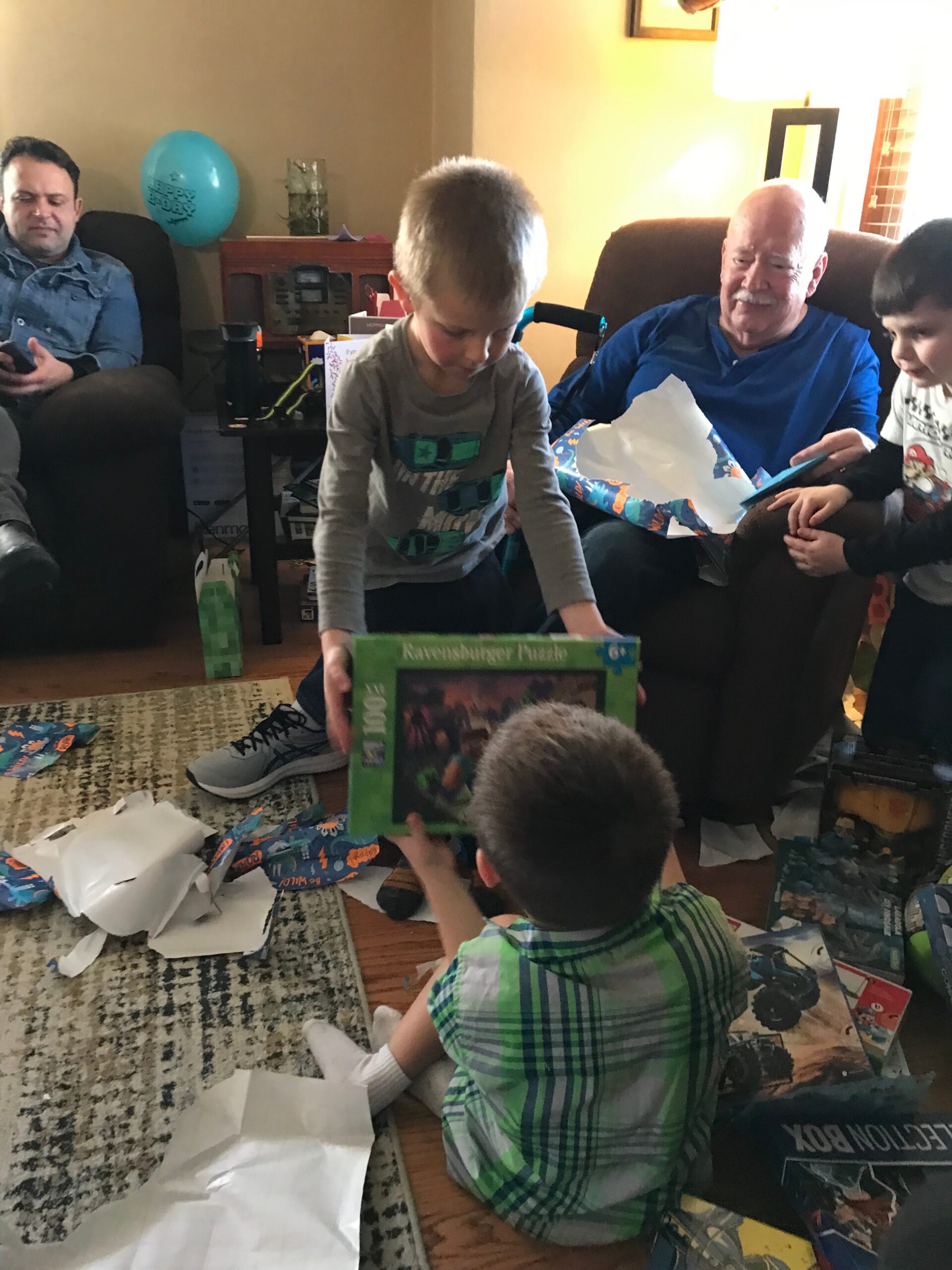 Young boy in gray shirt unwraps a gift while family sits nearby at a cozy living room birthday scene.