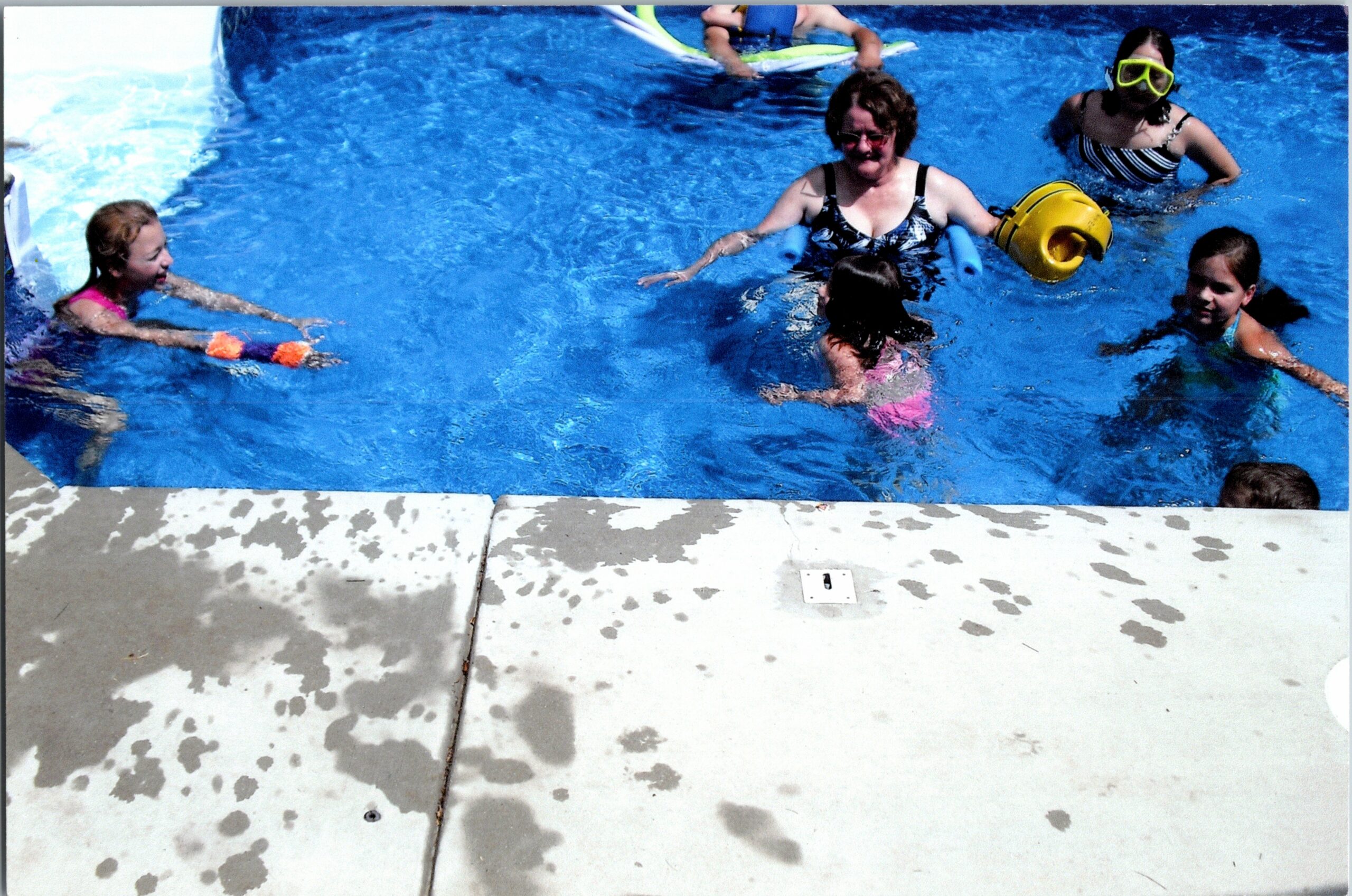 Several people, including children and an adult, swim in a bright blue pool near the edge, wearing swimsuits and flotation gear (yellow ring and foam noodles).