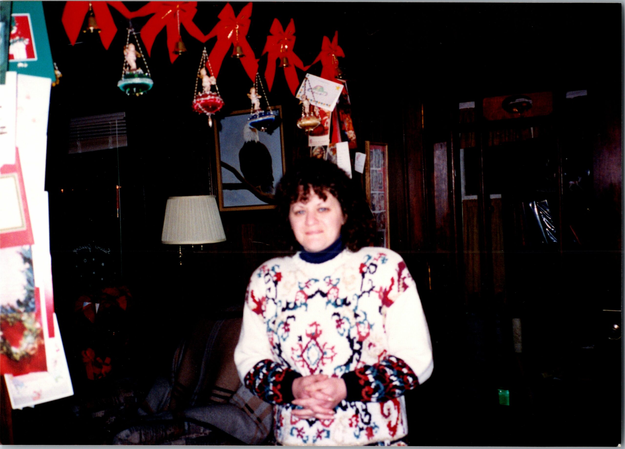 Woman with curly hair wearing a white Christmas sweater with red and blue patterns, standing indoors under red holiday garlands and ornaments.