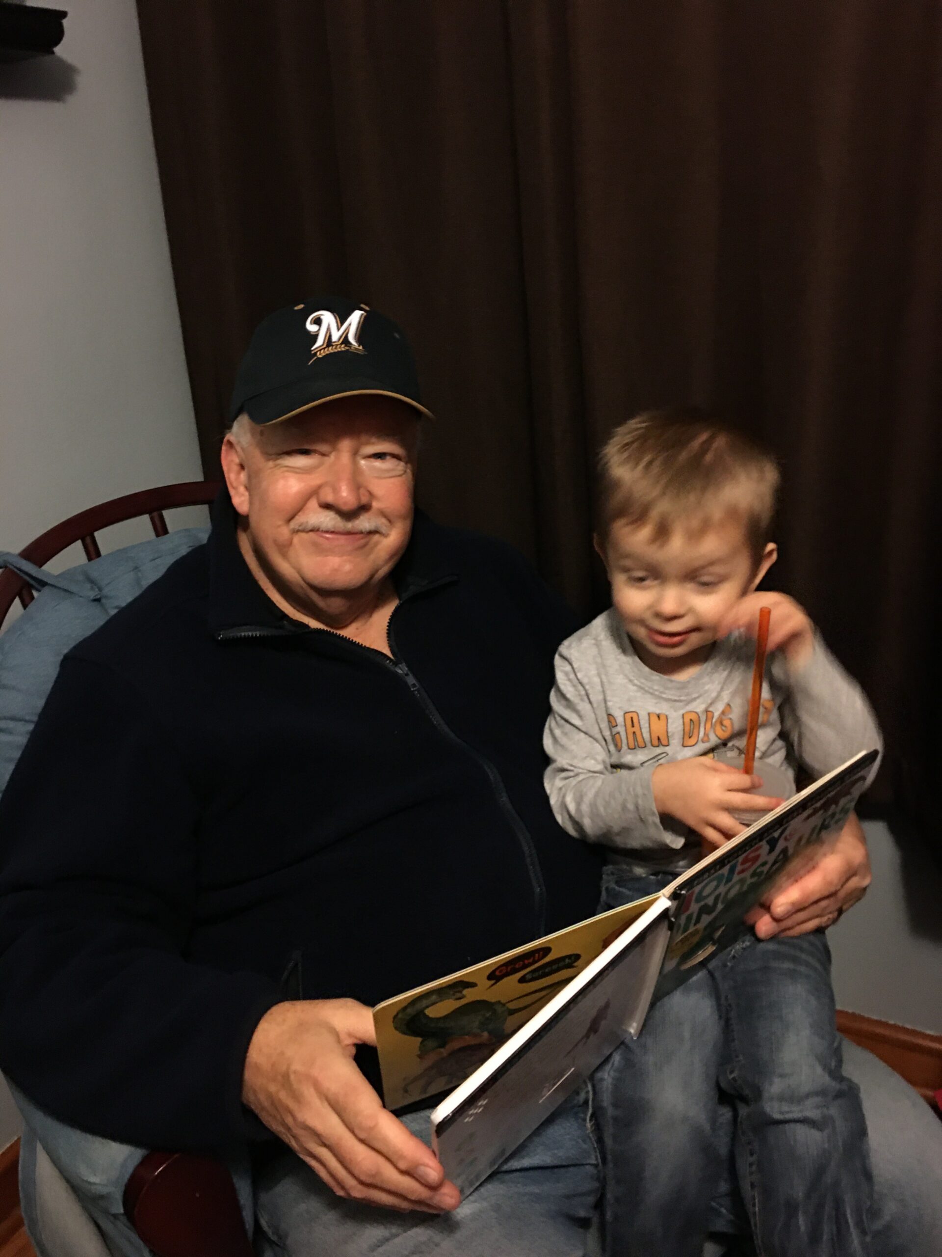 Older man with a mustache wearing a dark cap with an M logo, smiling as he reads a colorful children's book to a young boy beside him on a chair.
