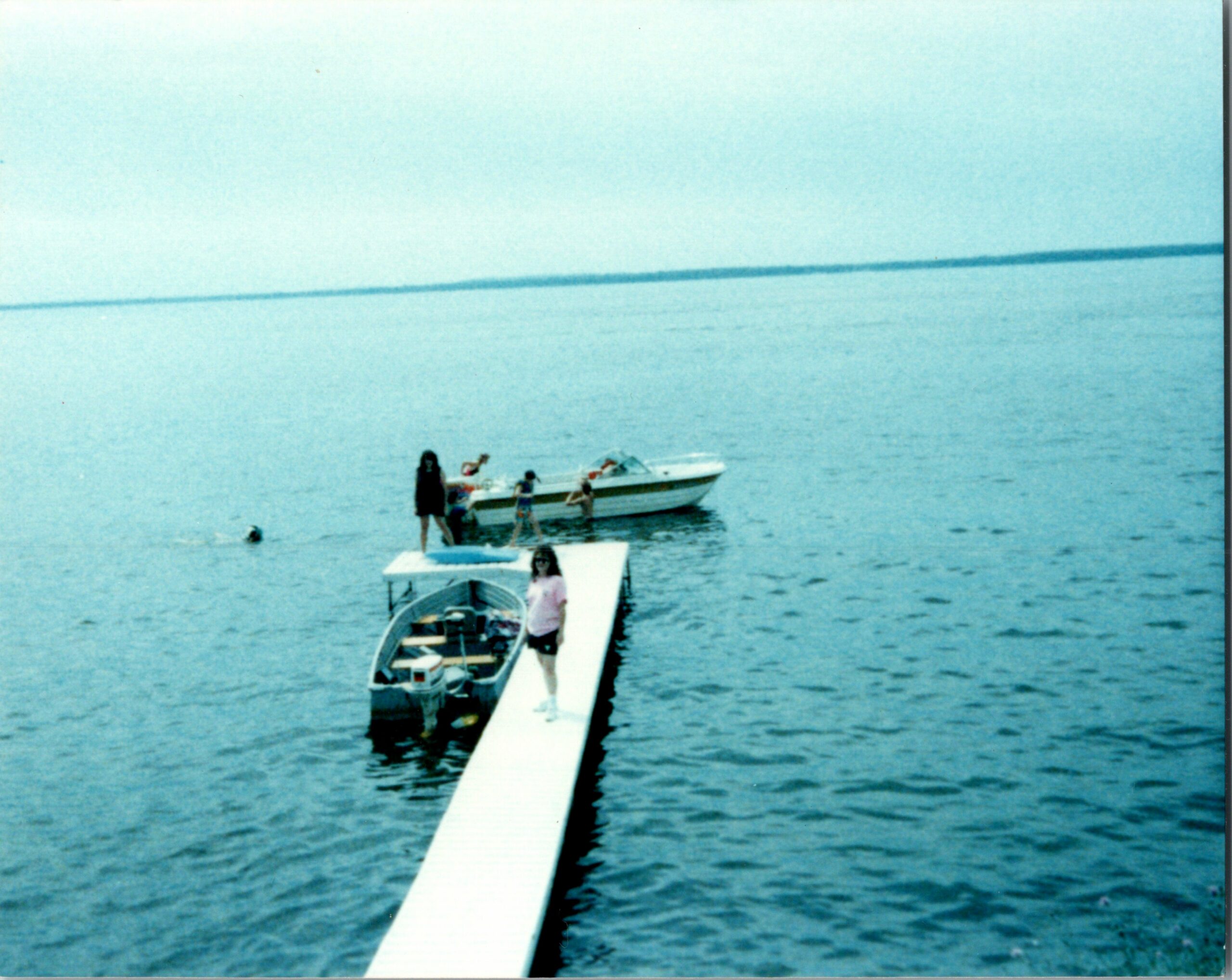 People stand on a white dock extending into calm blue water with small boats moored along the sides; a distant horizon line visible.