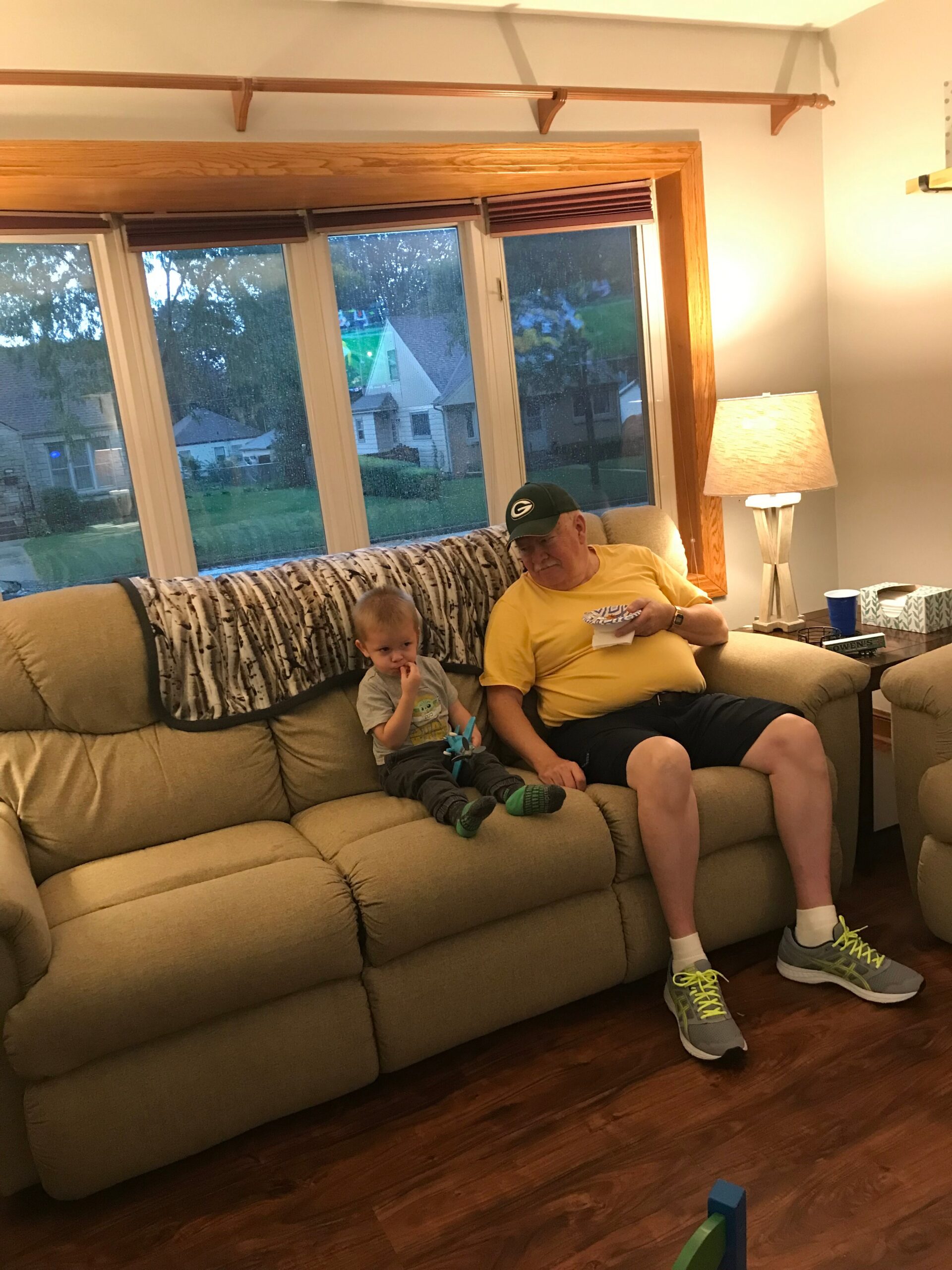 Older man in a yellow shirt and green cap sits on a beige couch with a small child, in a cozy living room with large windows and a lamp nearby.