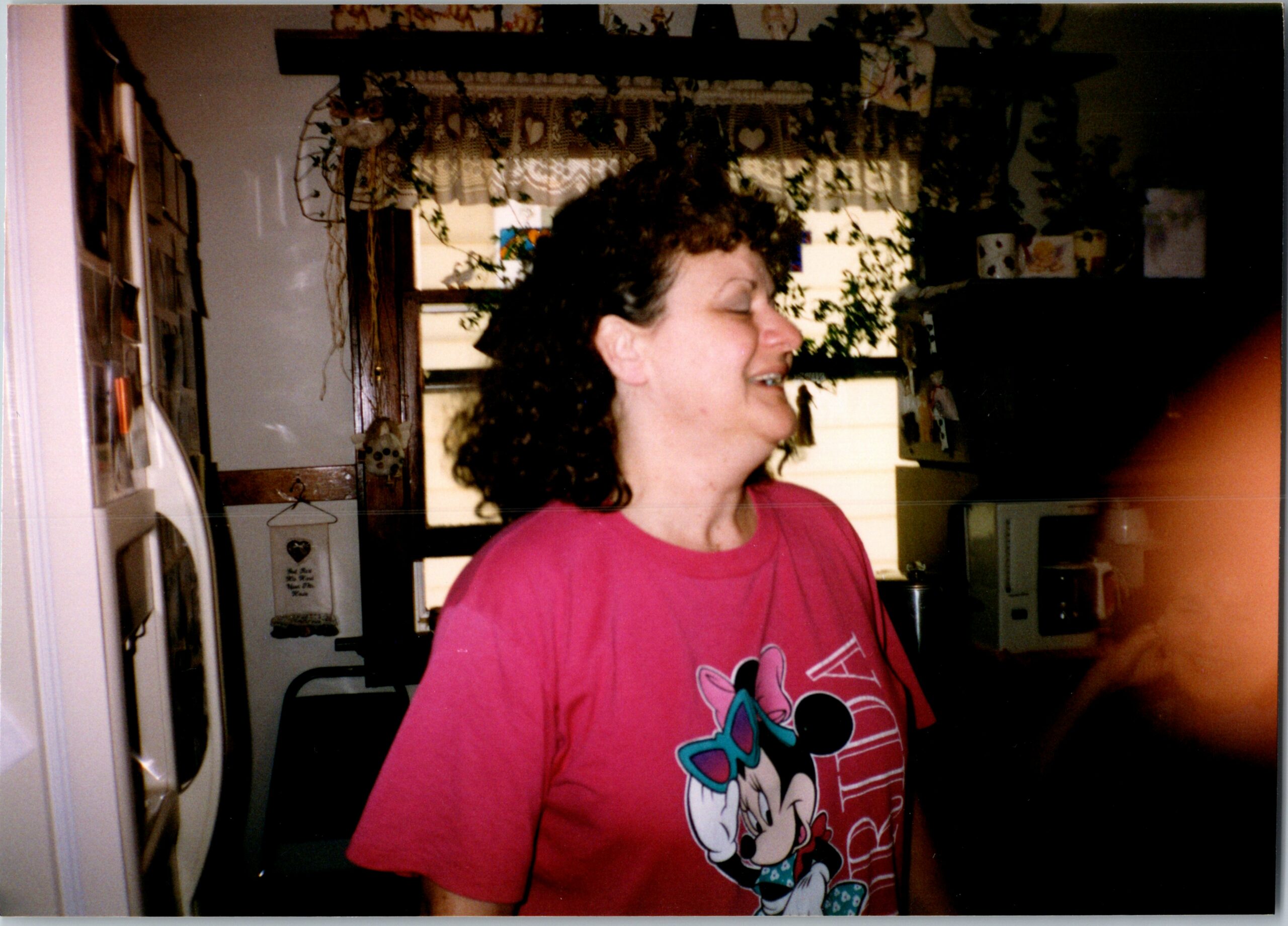 Side-profile of a woman with curly hair wearing a pink Minnie Mouse T-shirt, smiling in a cozy kitchen with hanging plants.