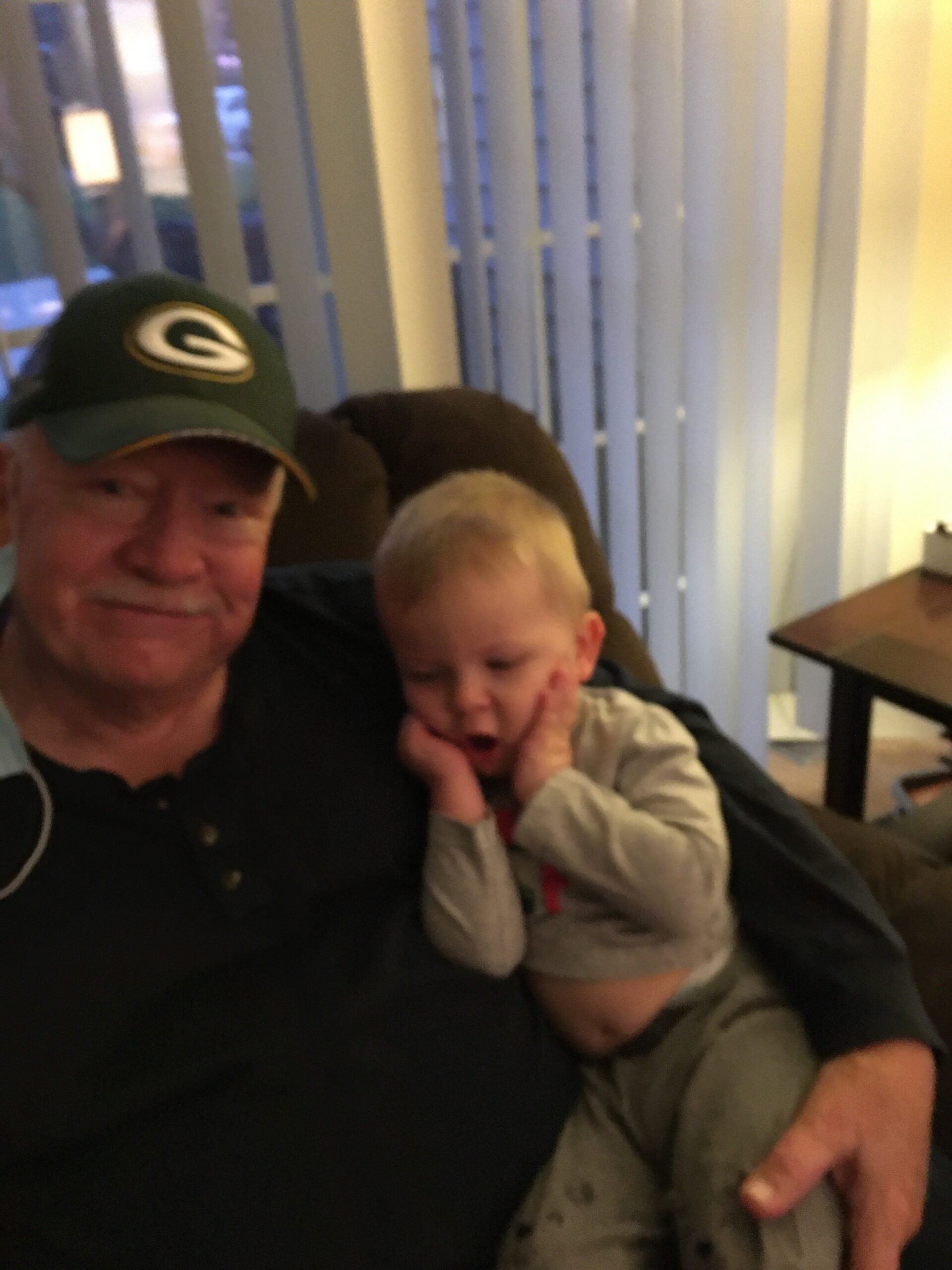 An older man wearing a green Packers cap sits on a dark couch, holding a young child who rests their head on the man's chest with a dirt-smudged face. Vertical blinds and a lamp are in the background.