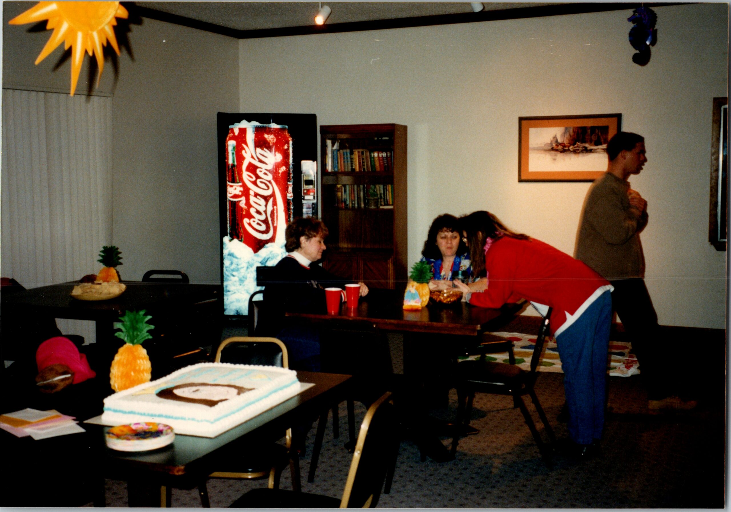 People at a casual party in a room with a large cake and pineapple decorations on a table; a Coca‑Cola vending machine, bookshelf, and wall art are in the background; one man stands to the right while others sit and chat.