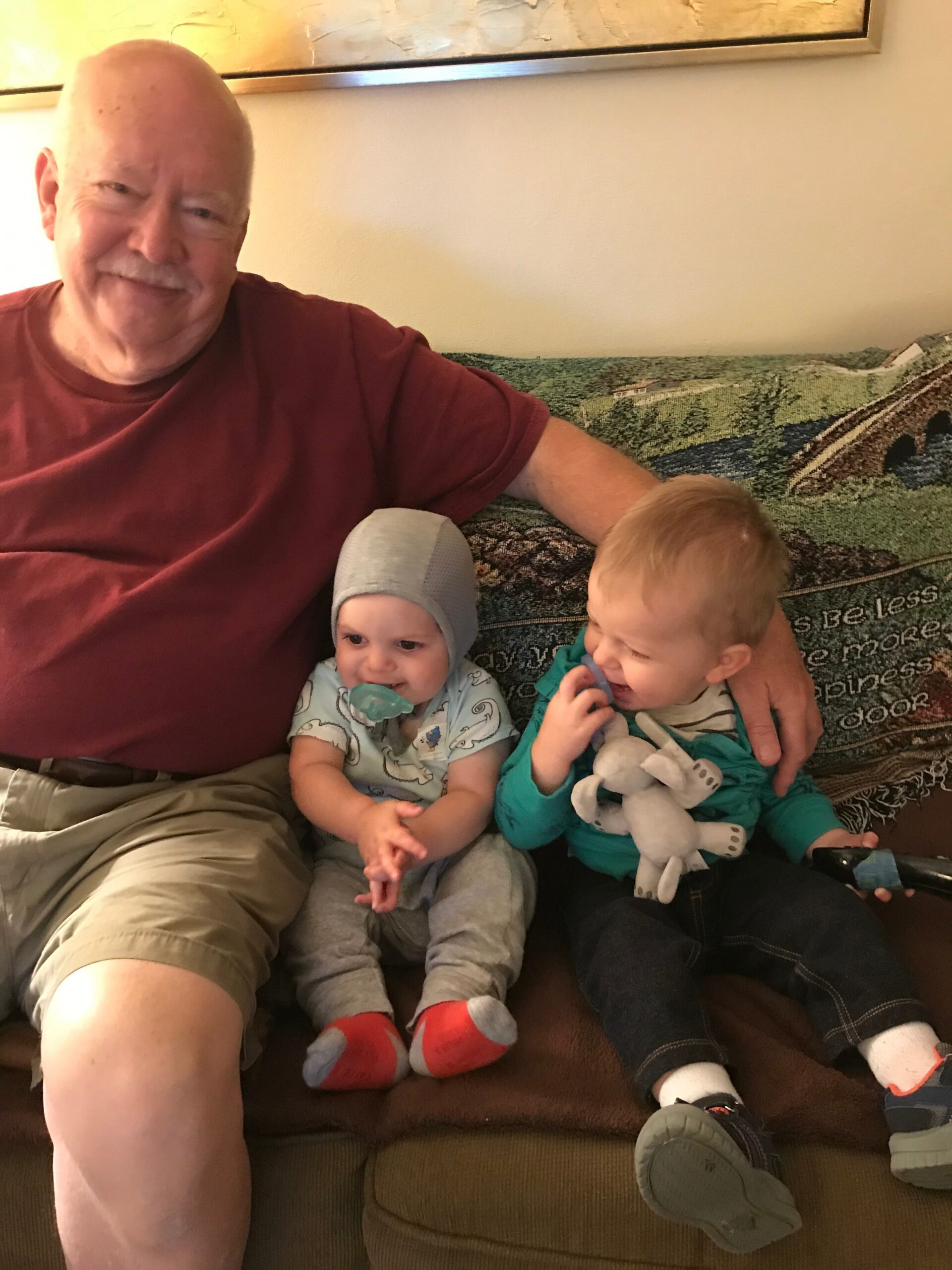 Older man in a red shirt sits on a couch with two toddlers, one with a pacifier in a gray hat and the other holding a stuffed toy on his lap.
