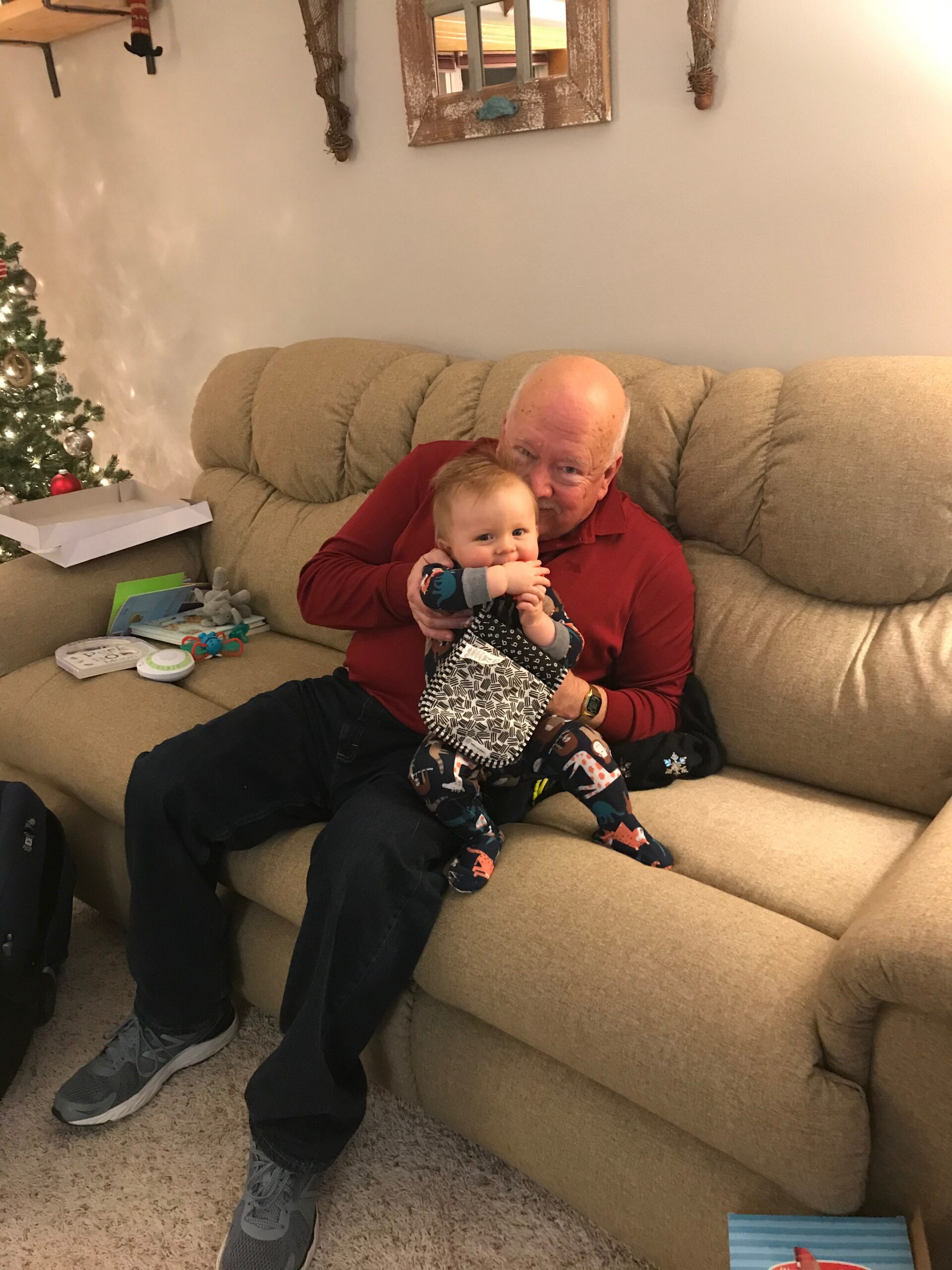 Grandfather in a red shirt sits on a beige sofa holding a smiling baby with a Christmas tree and gifts nearby.