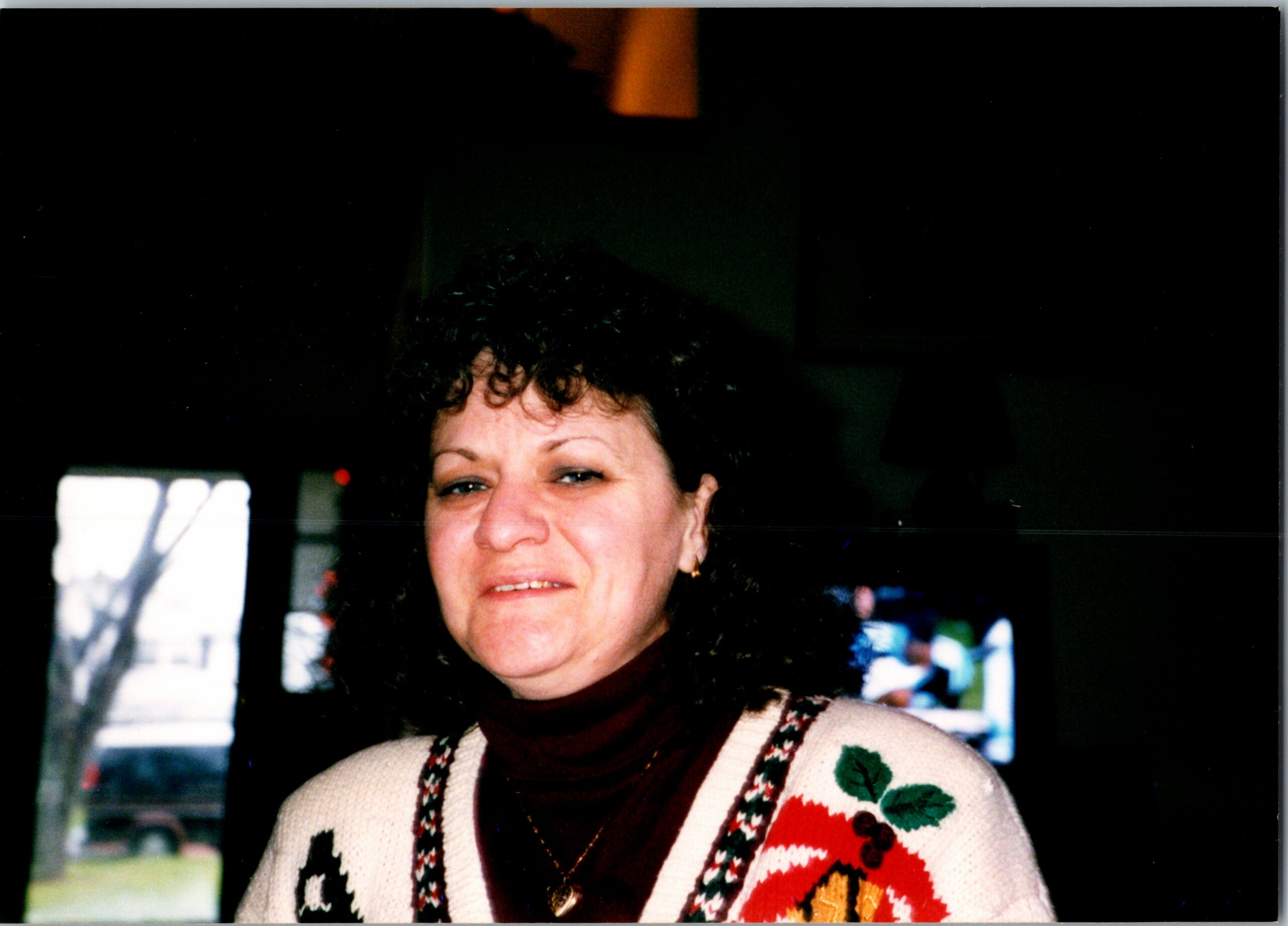 Smiling woman with short dark hair wearing a cream Christmas sweater with red and green patterns indoors.