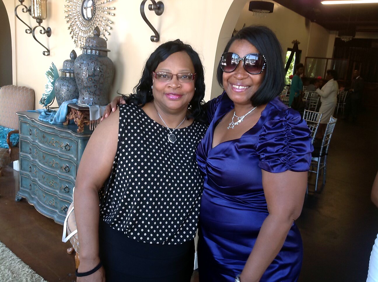 Two women posing together at an indoor social event; one in a black polka-dot top with glasses, the other in a royal blue dress wearing sunglasses, smiling.