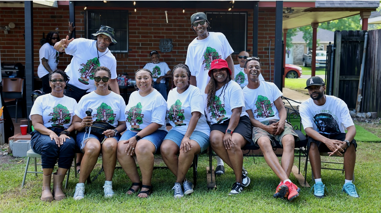 Group photo of ten people wearing matching white T-shirts with a tree graphic, seated on chairs in a backyard yard.