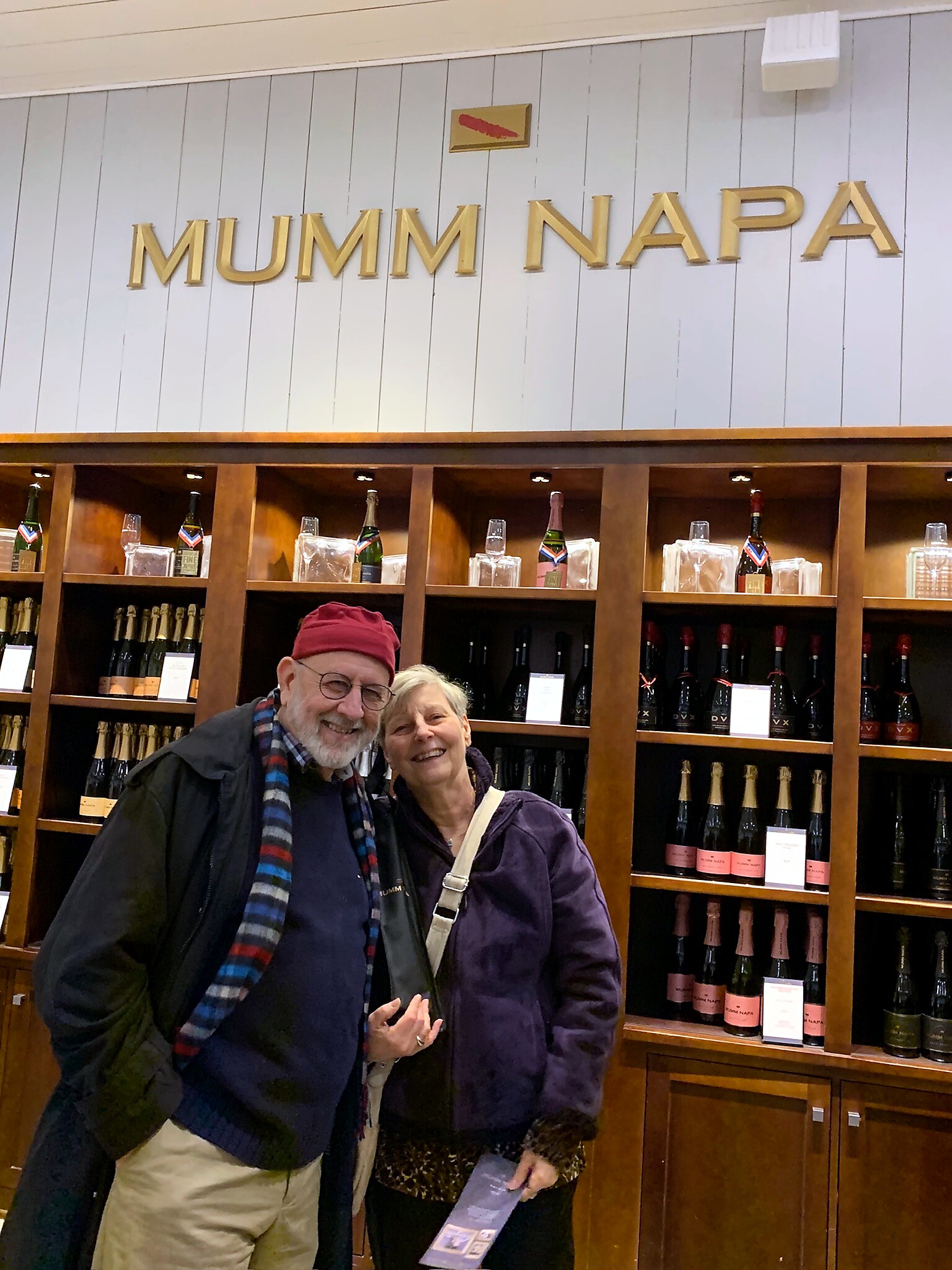 Couple posing and smiling in a wine shop with wooden shelves of bottles and a gold MUMM NAPA sign above them in Napa Valley. (informative image)