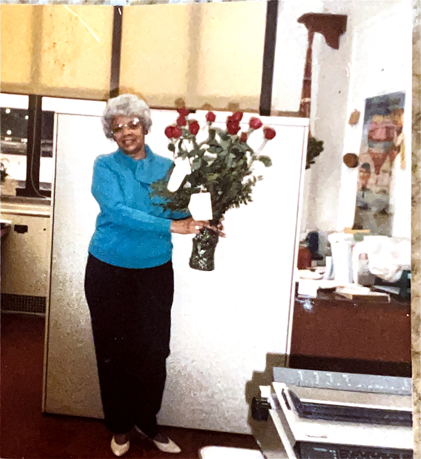 Woman with gray curly hair in a blue blouse holding red roses in a vase in a kitchen or room setting