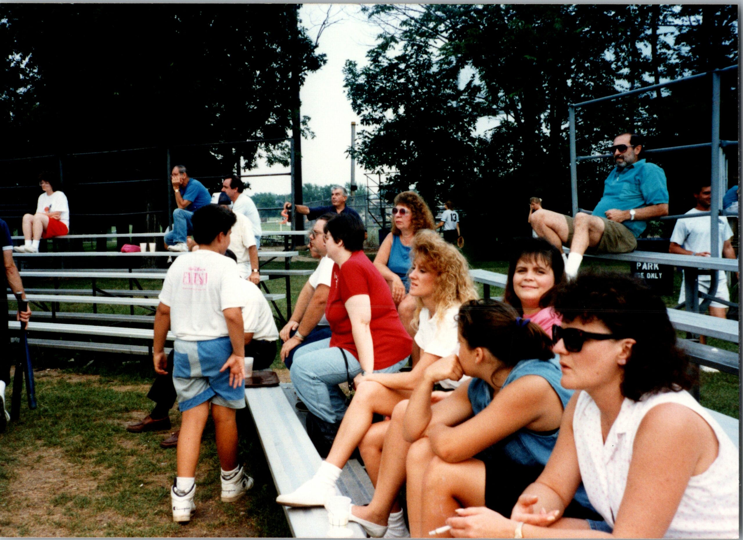 Spectators sit on metal bleachers at an outdoor park watching a game, with trees in the background and people chatting.