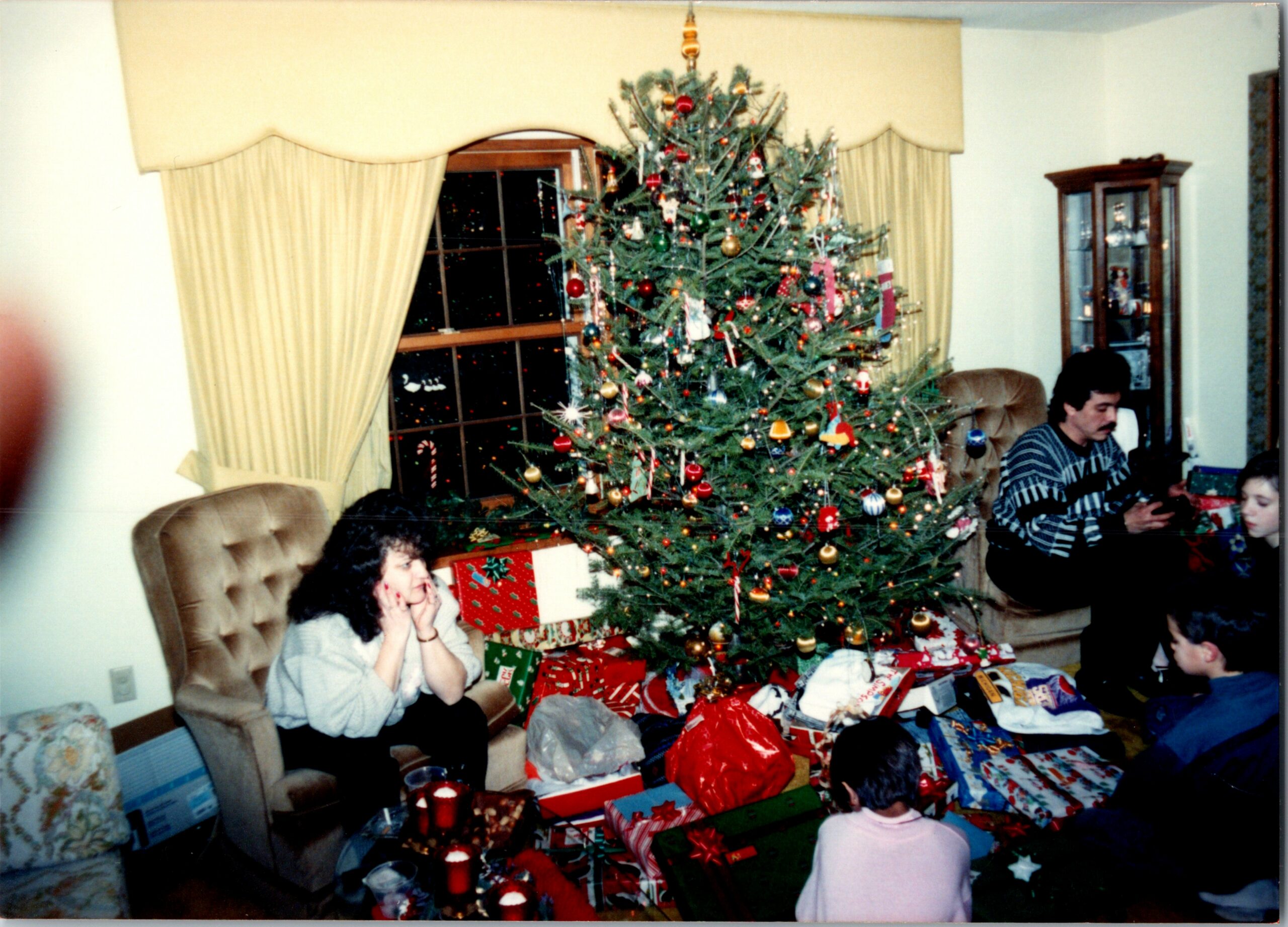 Cozy living room with a tall Christmas tree surrounded by wrapped gifts; a woman sits on a chair to the left and a man on the right.