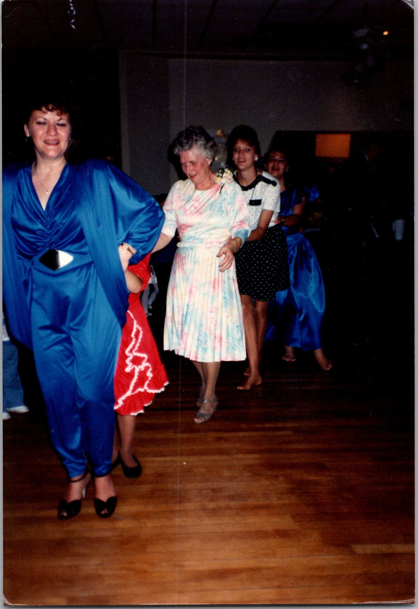 Women line-dancing at a formal event, wearing colorful dresses and heels, smiling as they move in a row on a wooden floor.