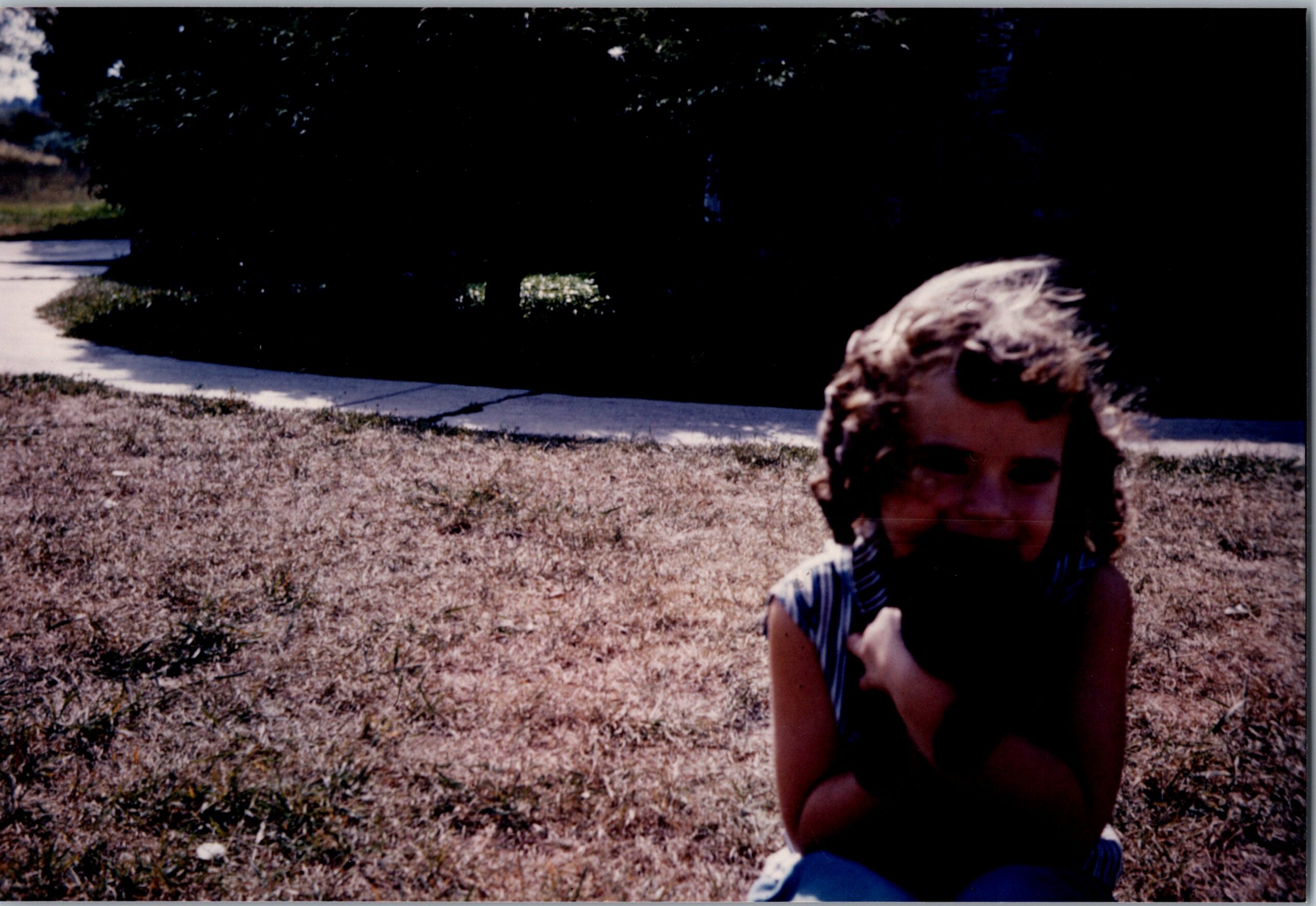 Young girl sitting on dry grass beside a sidewalk with bushes in the background on a sunny day.