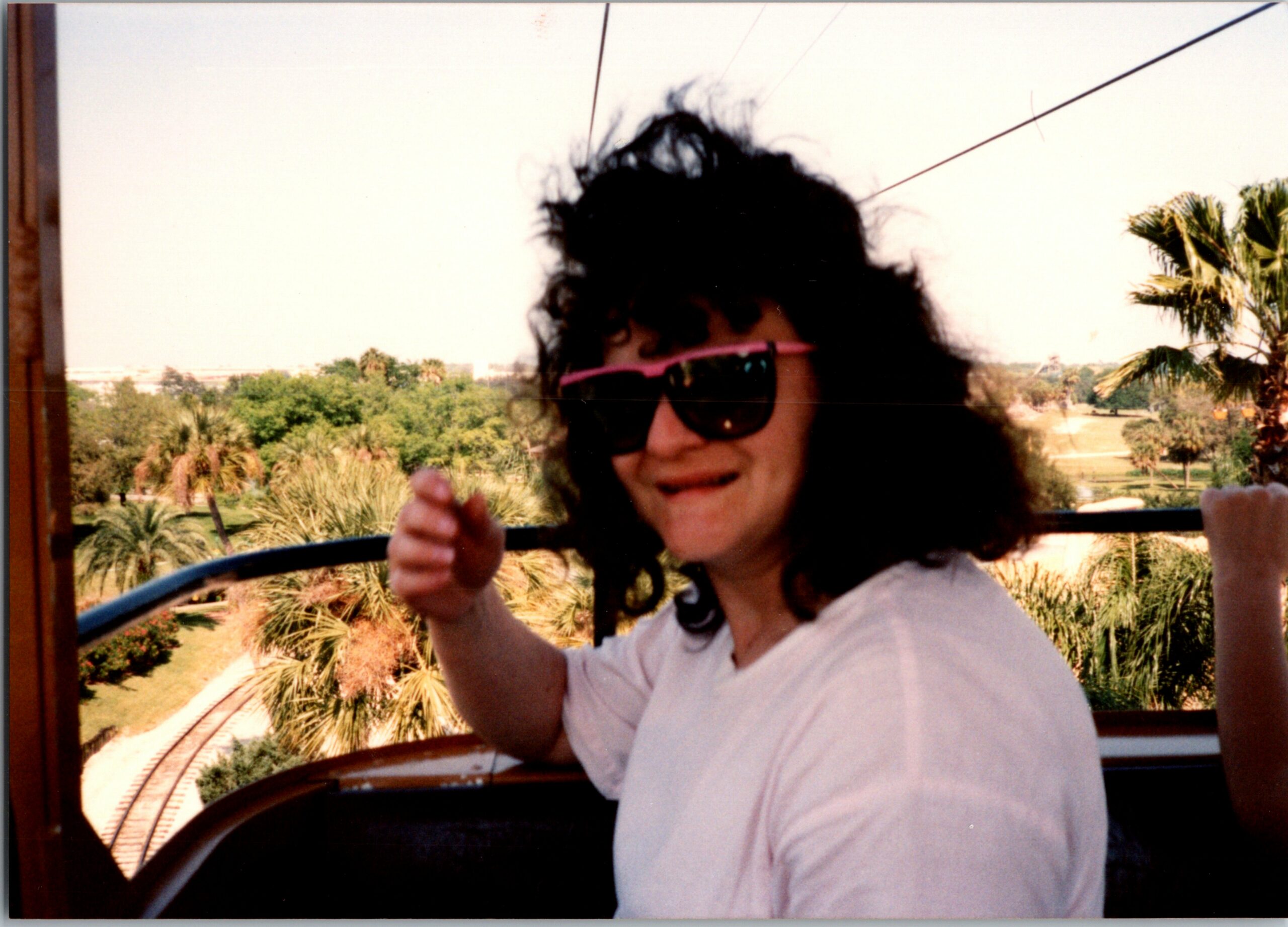 Child with curly hair wearing pink sunglasses giving a thumbs-up on a sunny outdoor deck with palm trees in the background.