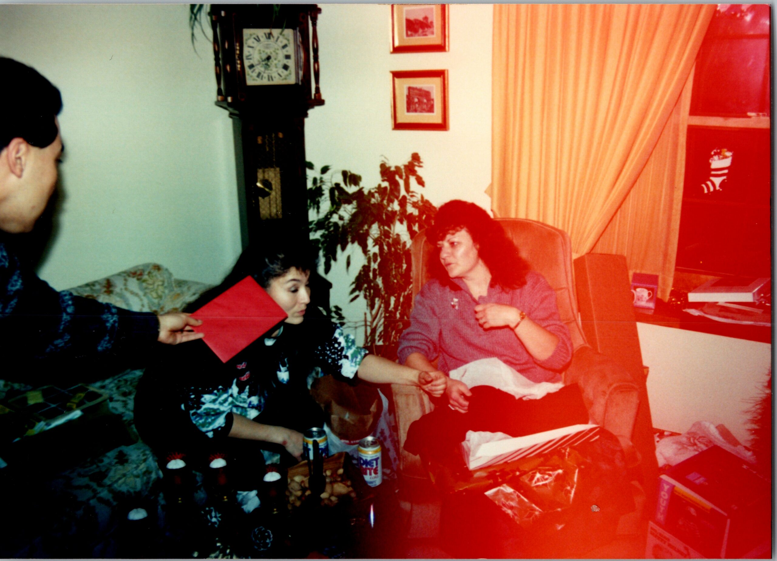 Family in a living room sharing a red envelope; a woman sits on a chair while a child hands her a red envelope, with cupcakes and drinks on the table and a tall clock against the wall nearby.