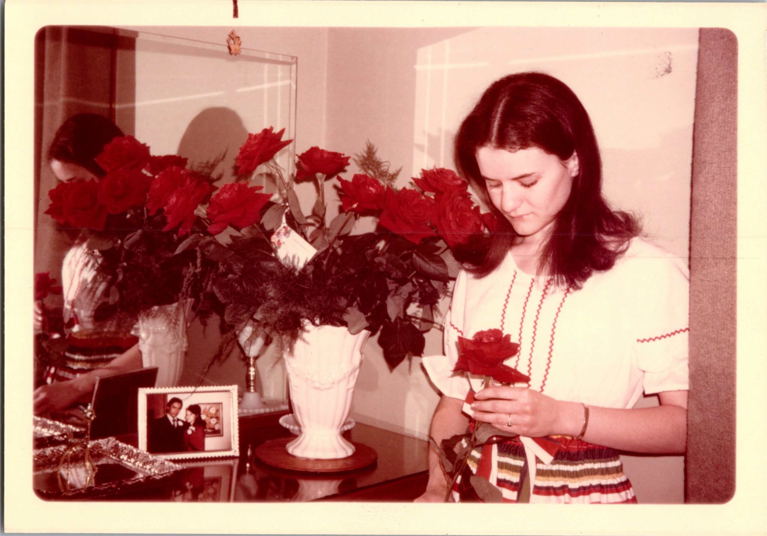 Woman in a striped dress stands beside a vase of red roses on a dresser, looking down at something in her hands.