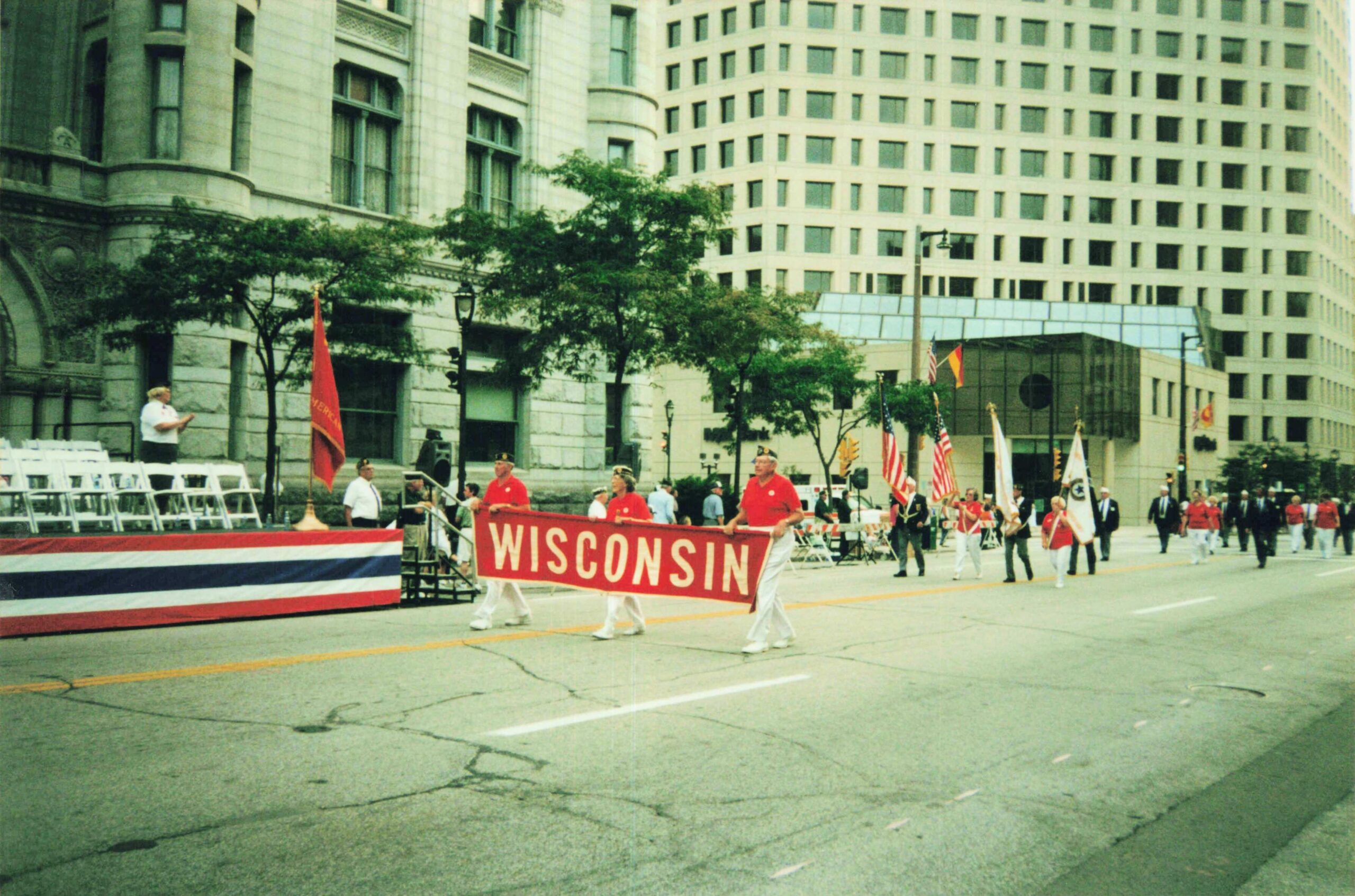 Parade marchers hold a red Wisconsin banner along a city street with flags and spectators lined up on the curb.