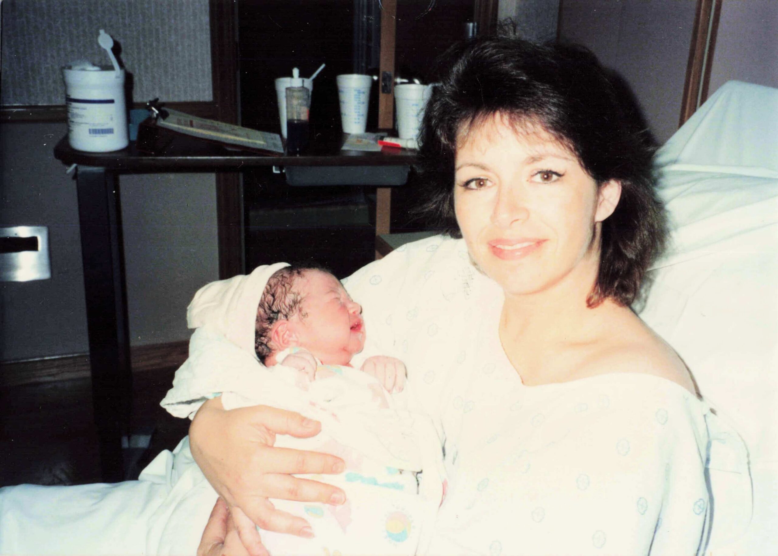 Woman in a hospital gown cradling a newborn baby in a hospital room with medical supplies in the background.