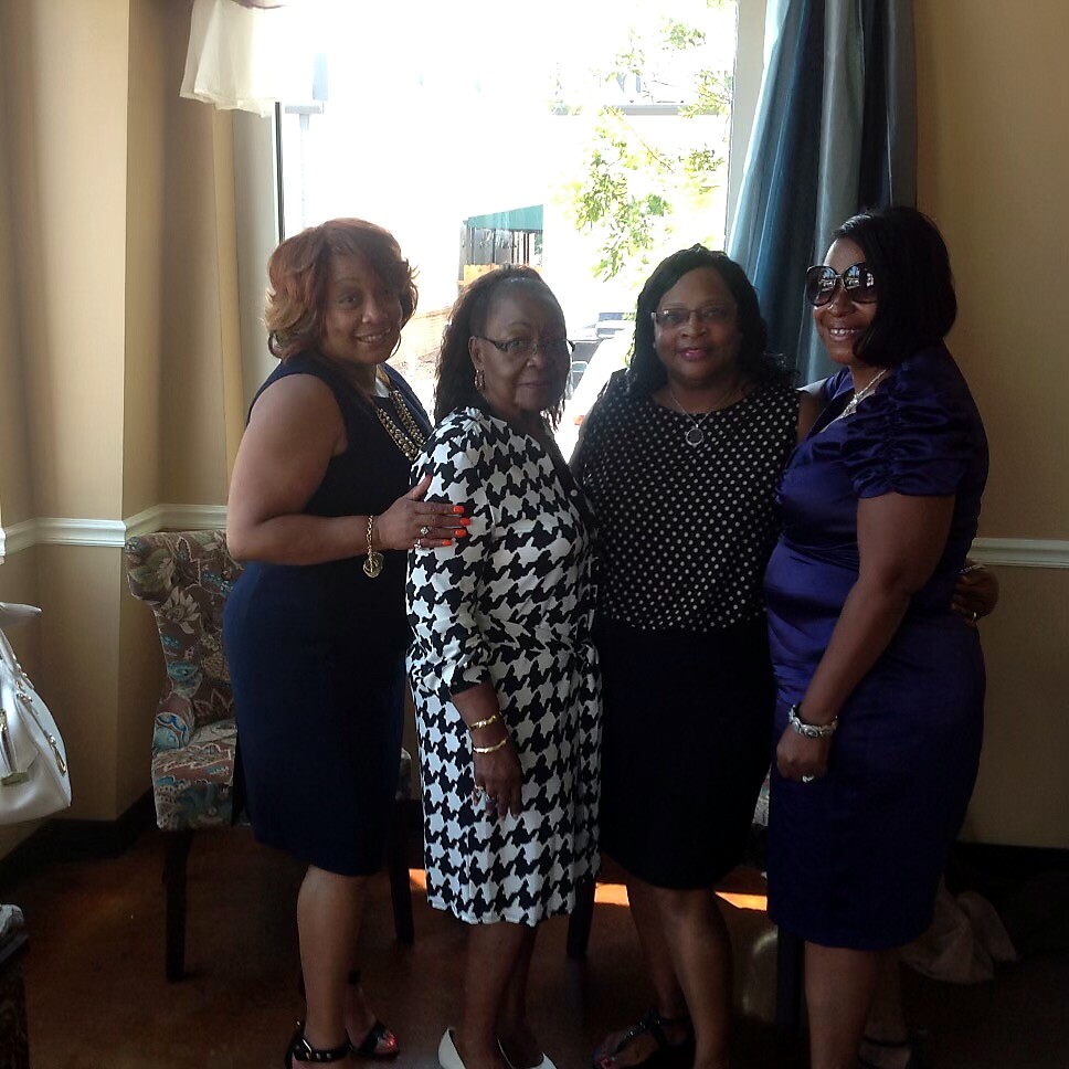 Four women standing together indoors, posing for a group photo and smiling.