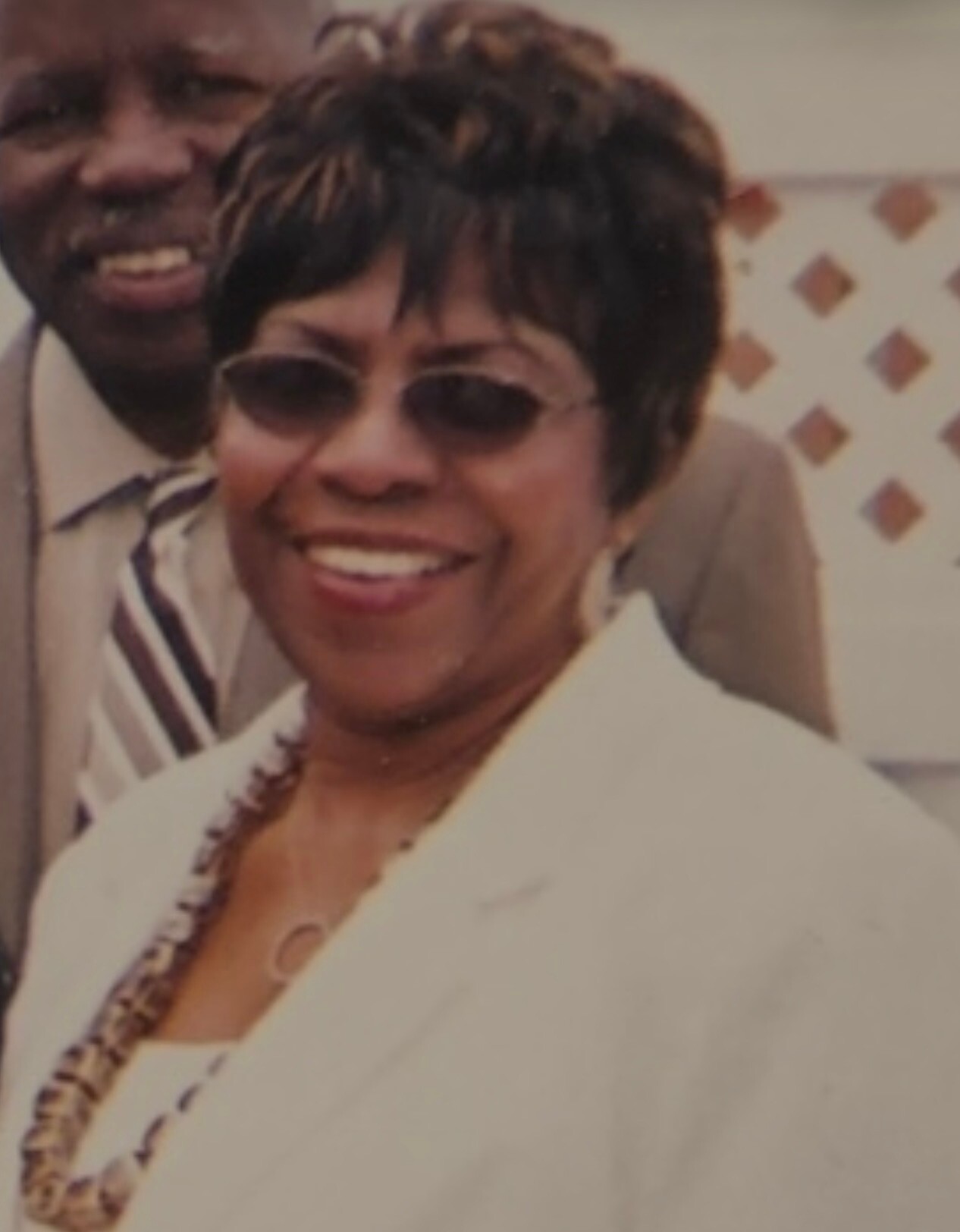 Smiling African American woman wearing sunglasses and a white blazer, with a beaded necklace visible.