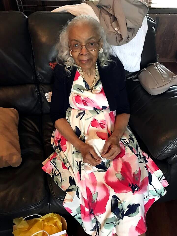 Older woman with gray hair and glasses sits on a black leather couch in a floral pink-and-white dress, holding a napkin.