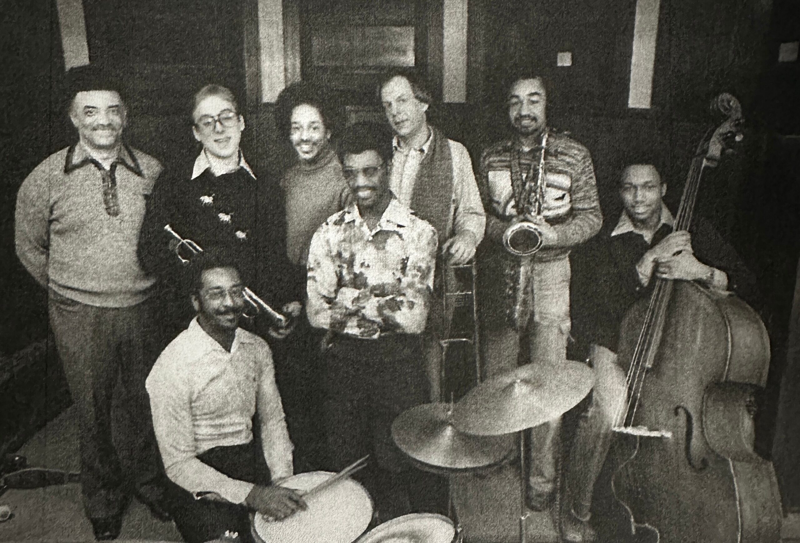 Black-and-white photo of a multi-person jazz band posing together with their instruments (guitar, saxophone, trumpet, bass) in a dim room, mid-20th century style attire.